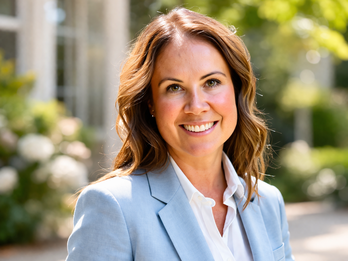 A smiling woman with brown wavy hair wearing a light blue blazer and white shirt, outdoors with greenery in the background.