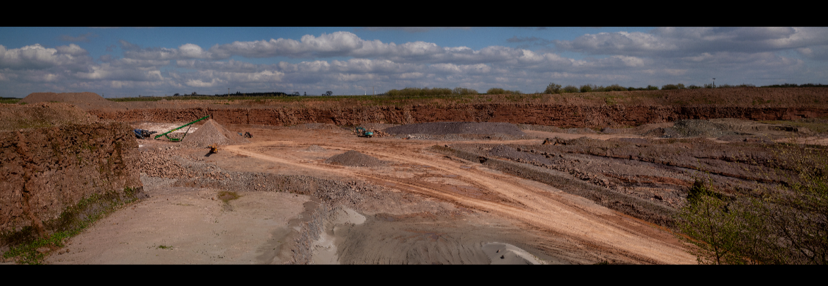 Open-pit mining site with excavator and trucks, dirt roads, and exposed earth layers under a partly cloudy sky.