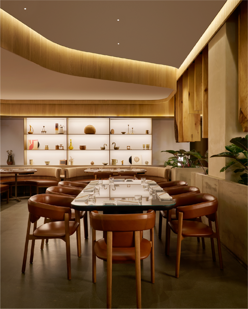 Interior of a modern restaurant with wooden accents, a marble table with glasses and cutlery, brown leather chairs, and decorative shelves with pottery and art pieces.