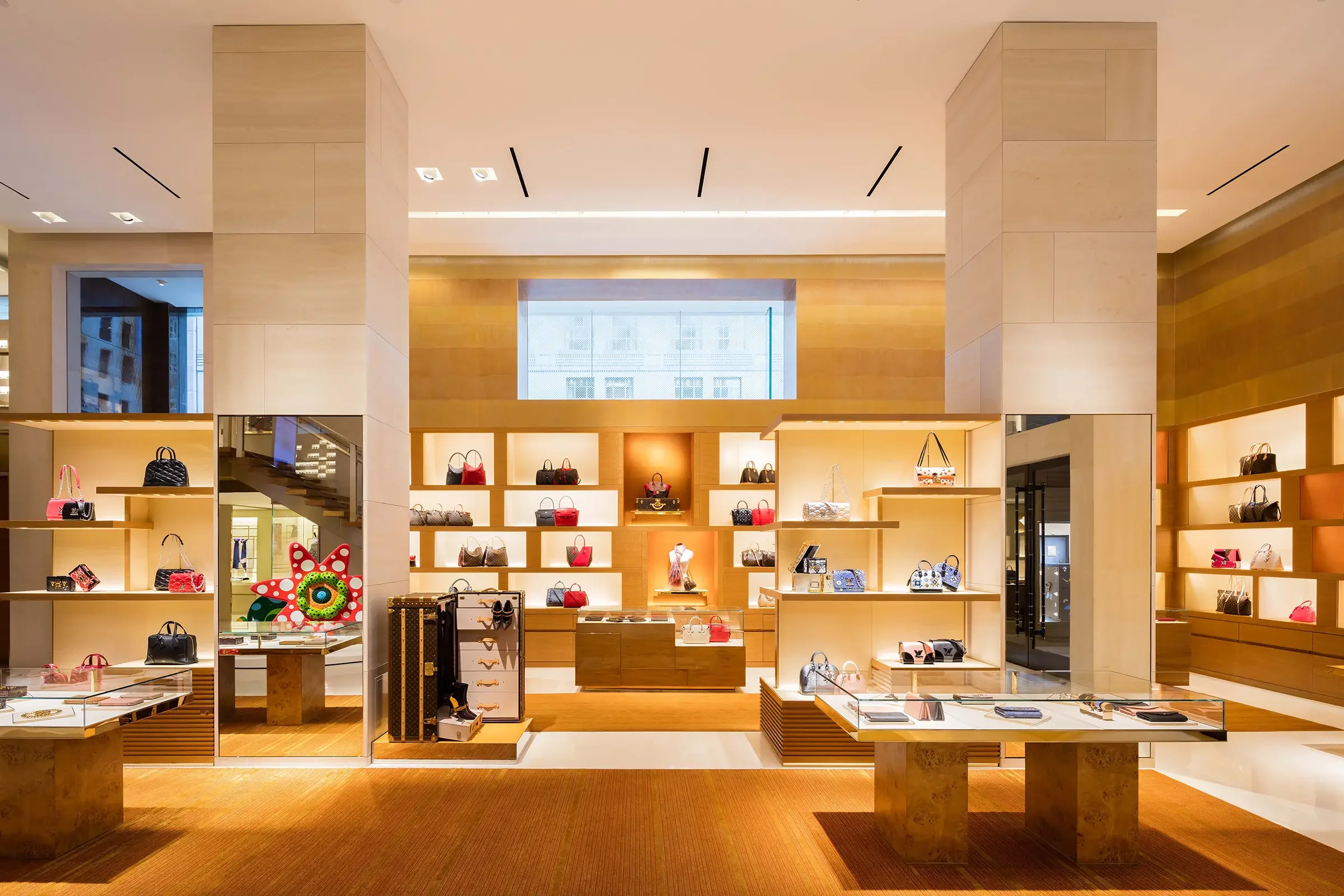 Inside a luxury handbags store with display shelves holding various designer handbags, a colorful plush flower toy, and a glass display table with accessories.