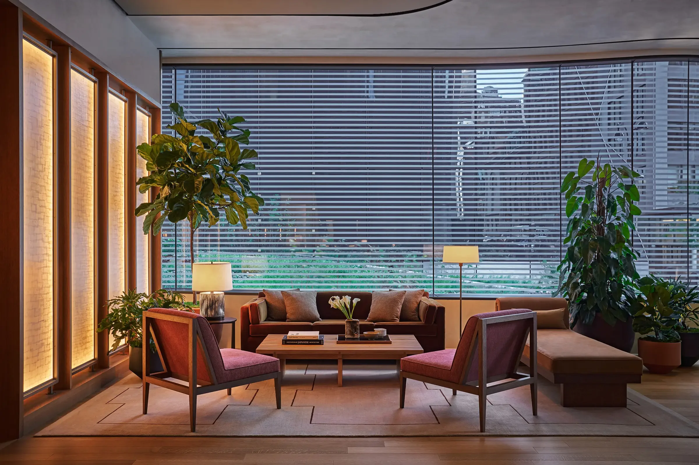 Modern living room with large window blinds, indoor plants, sofas, armchairs, lamps, and a coffee table with books and flowers.