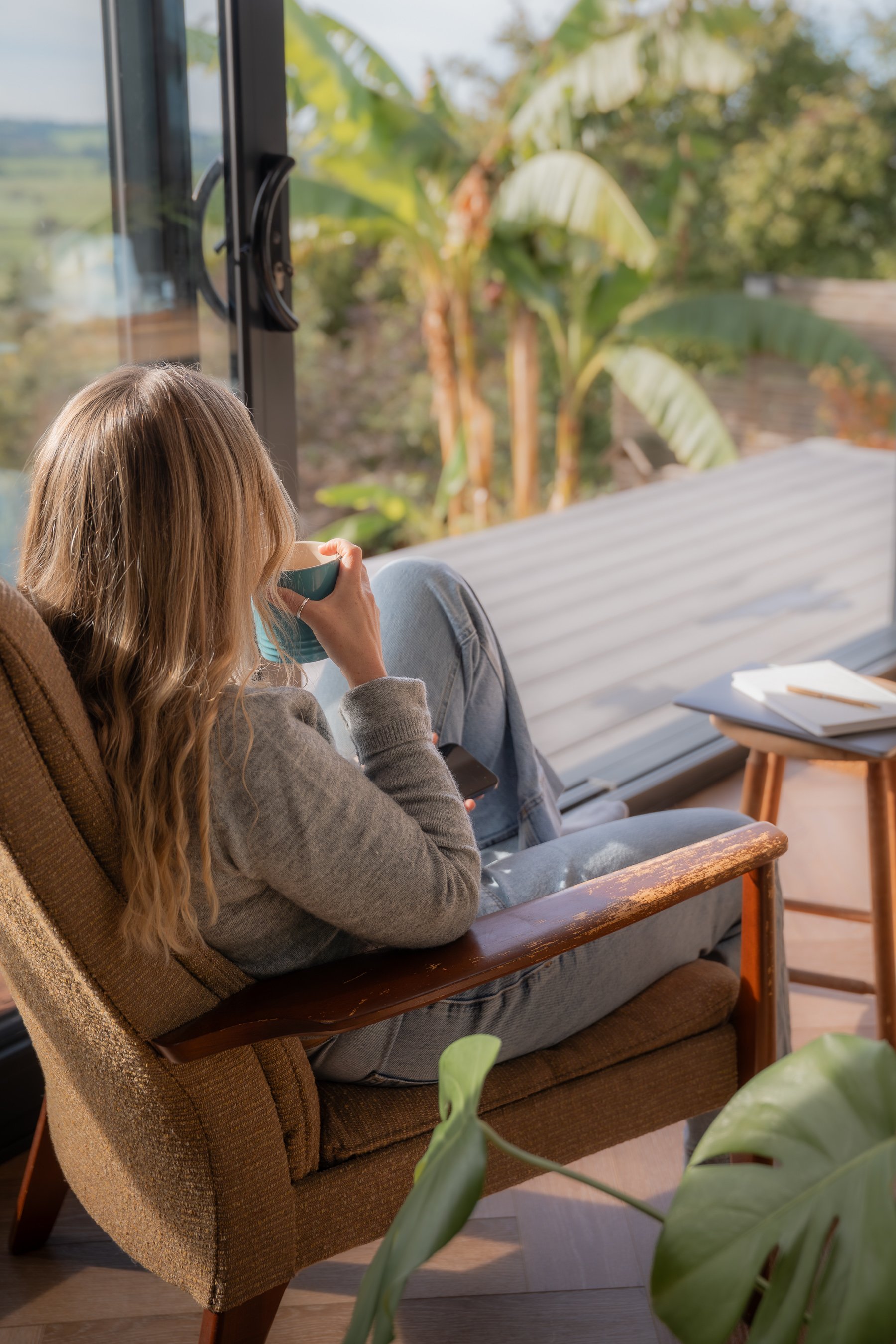 A woman with long blonde hair sitting in a brown armchair by a large window, drinking from a mug, with green plants and trees outside.