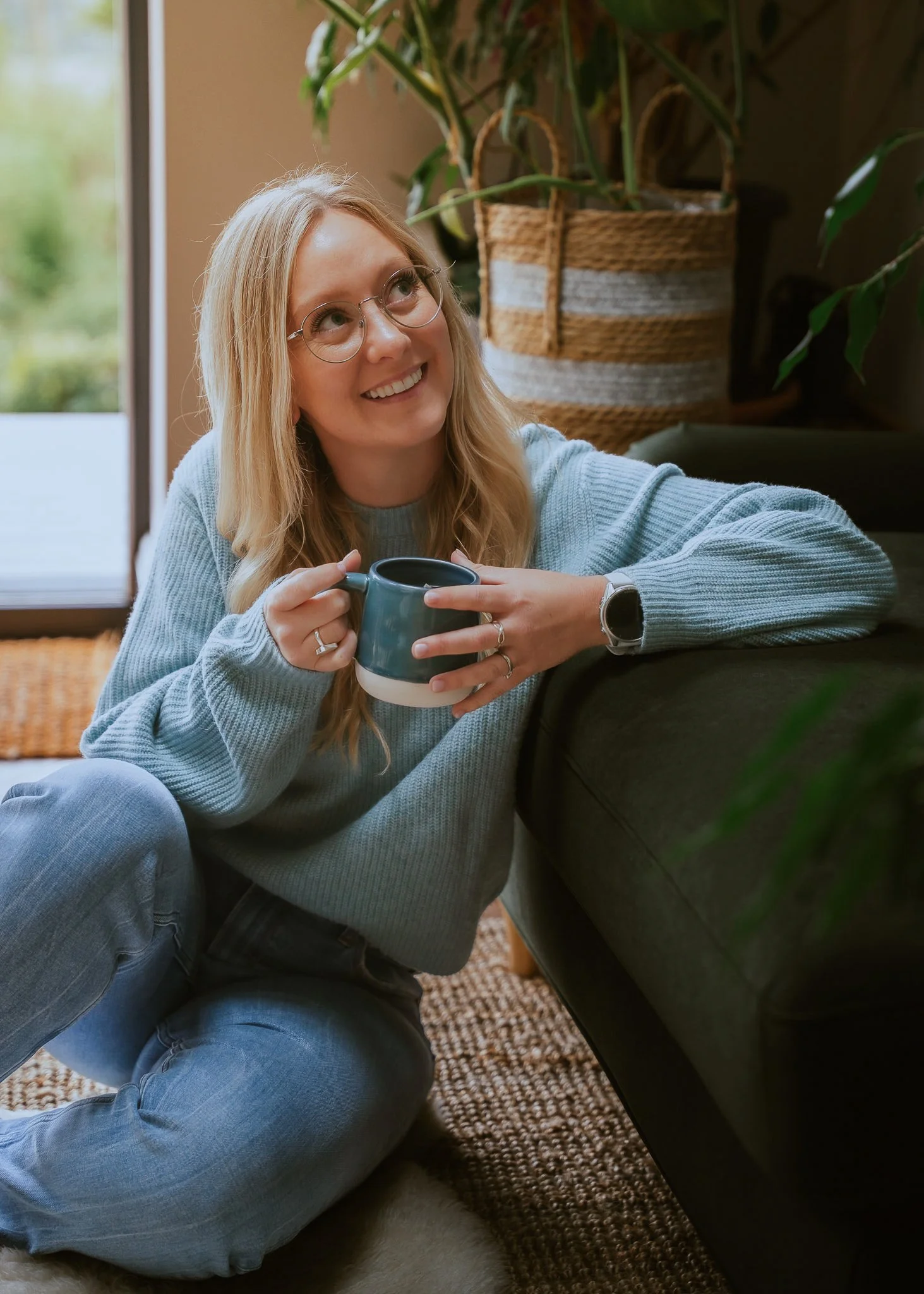 A woman with blonde hair and glasses sitting on the floor, smiling and holding a blue mug, in a cozy room with a window, a large plant in a woven basket, and a patterned rug.