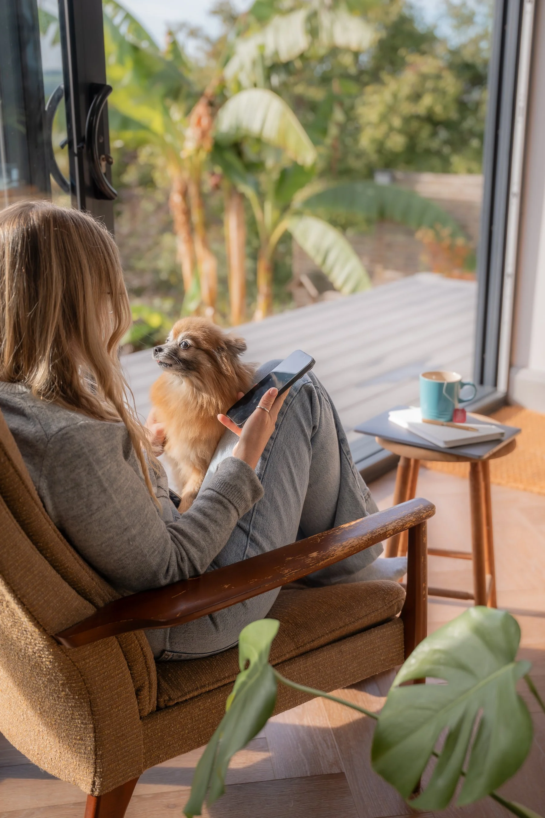 A woman sitting on a brown armchair indoors, holding a small brown dog and a smartphone, looking out a large window at green plants outside. There is a wooden table table beside her with a mug, a notebook, and a pen.