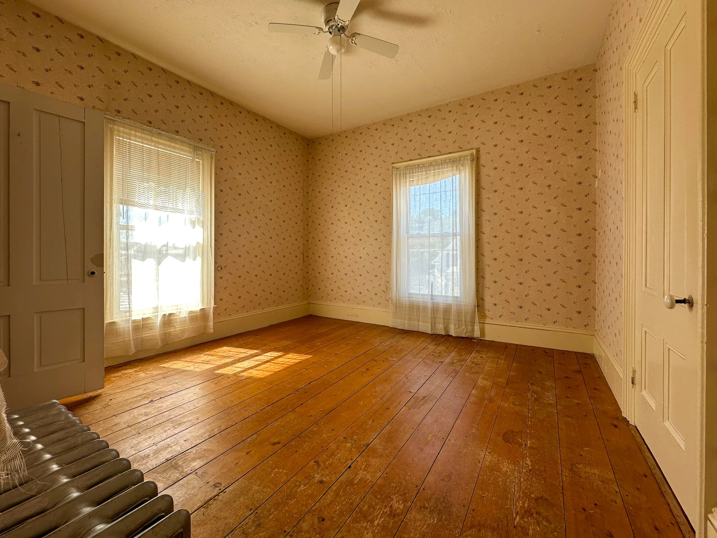Empty room with wooden floor, wallpapered walls, two windows with lace curtains, ceiling fan, and a radiator in the corner.