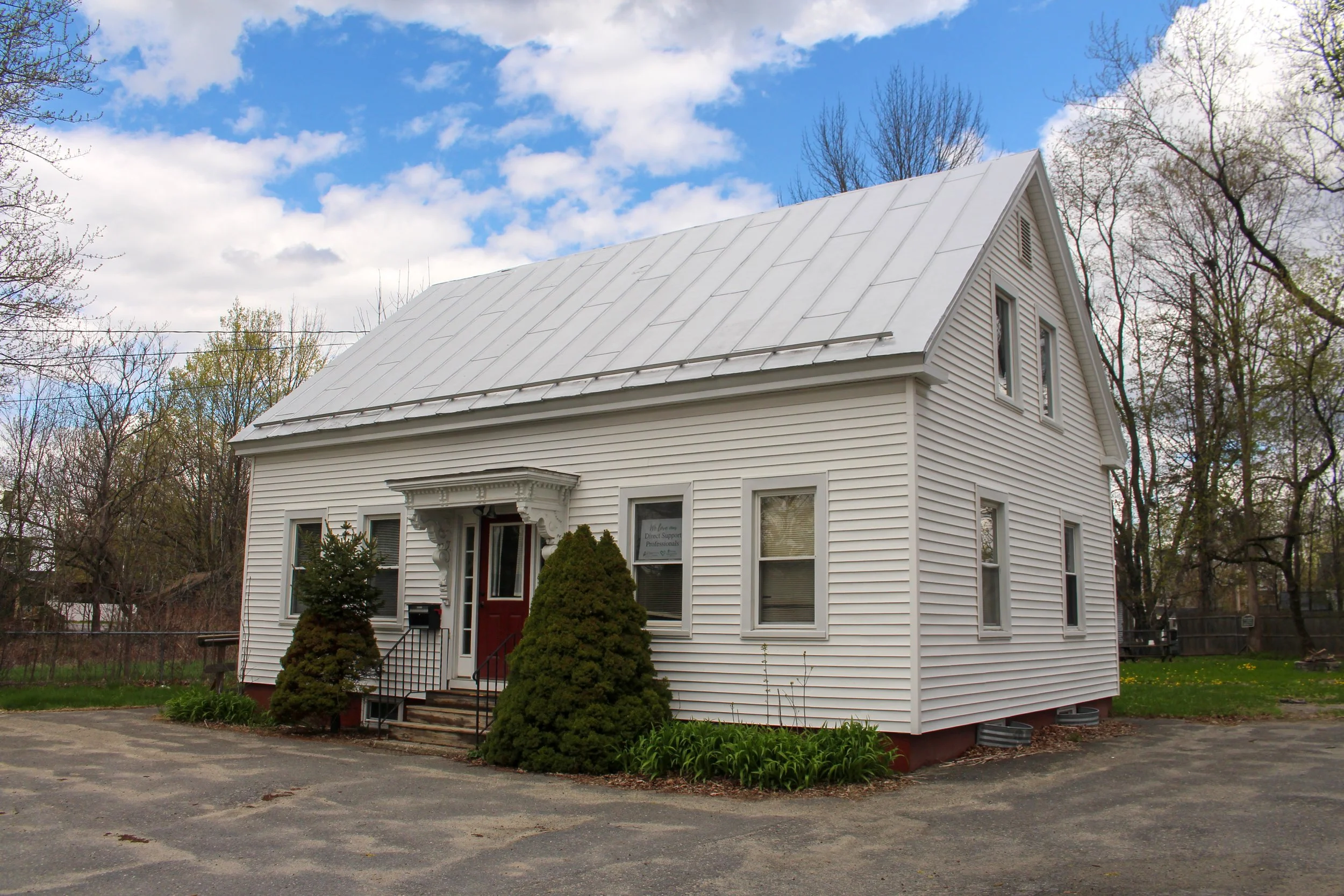 White, two-story house with a metal roof and a small front porch with stairs, surrounded by trees and bushes, under a partly cloudy sky.