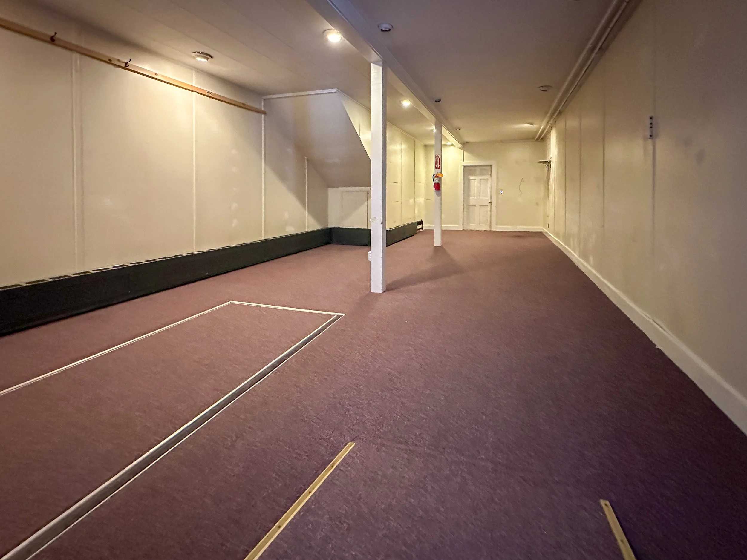 An empty room with beige walls, a purple carpet, and a white door in the background. There are two vertical support columns and a strip of molding along the ceiling with built-in lighting. Some yellow masking tape is visible on the carpet, and a fire
