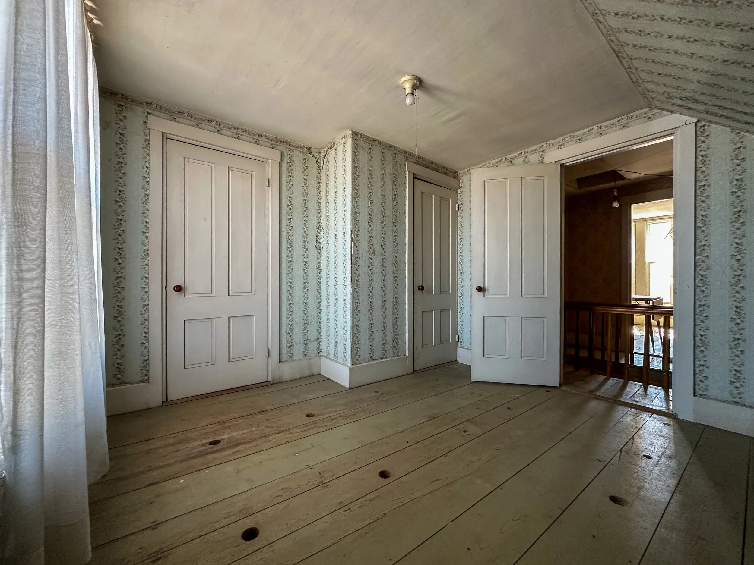 An empty room with wooden floors, floral wallpaper, two closed closet doors, and an open doorway leading to another room with a window and a hanging light bulb.