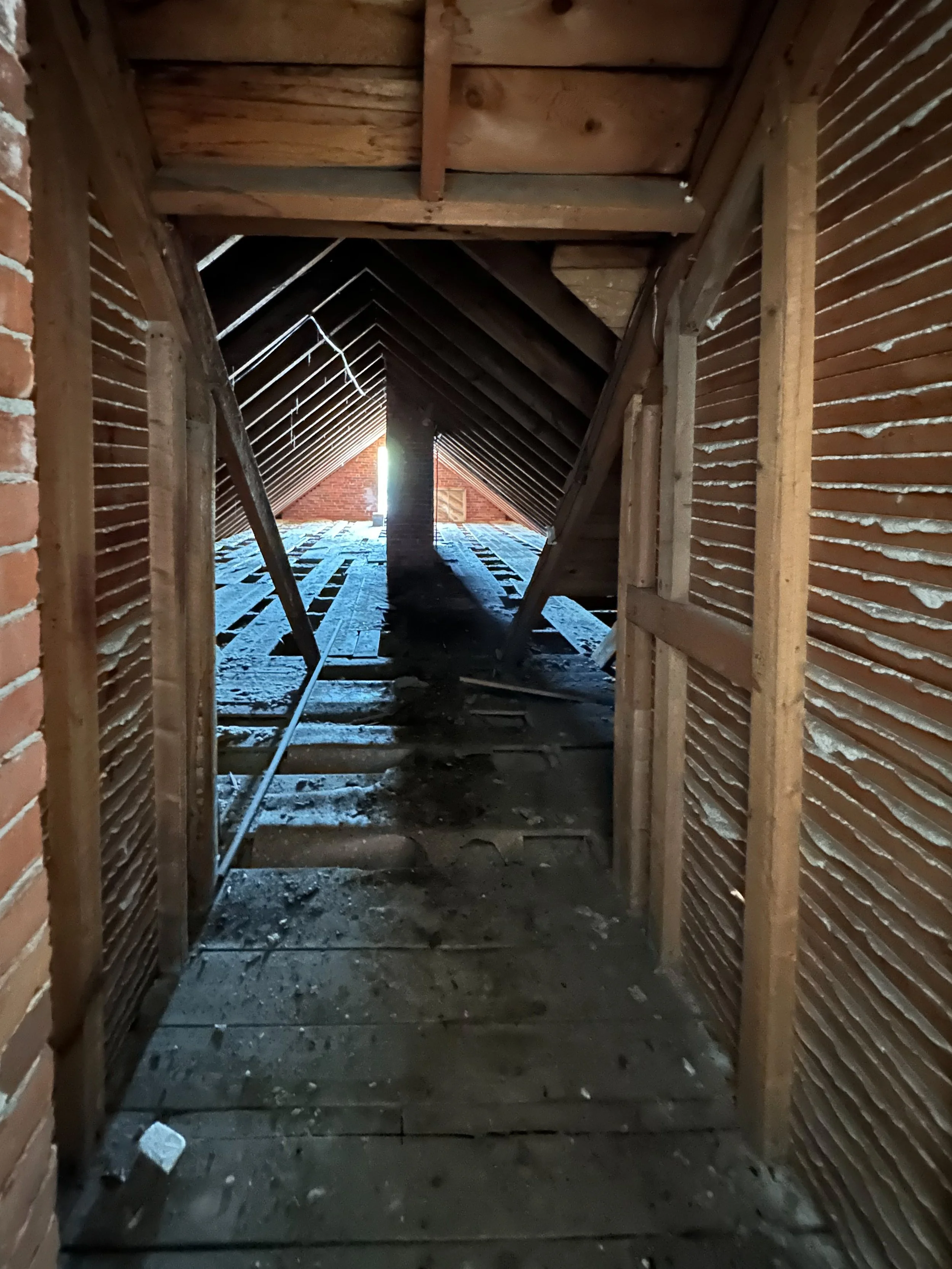 The inside of an attic space under construction, showing wooden beams, brick walls, and a ladder, with sunlight coming through an opening.