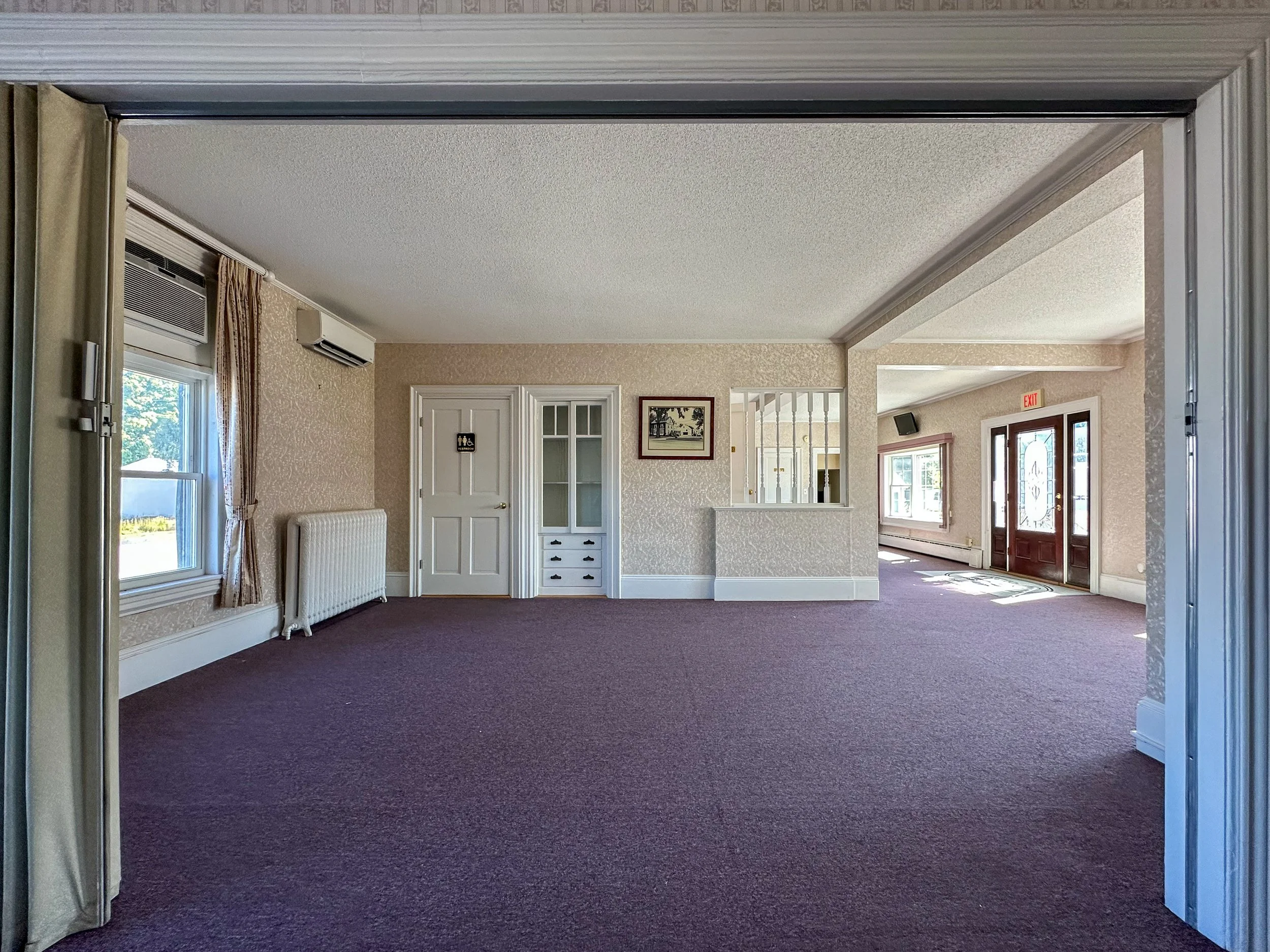 Interior view of a room with beige textured wallpaper, a black carpet, and windows with curtains. There is a white radiator, a door with a sign indicating restroom, and a framed picture on the wall. Part of a doorframe is visible in the foreground.