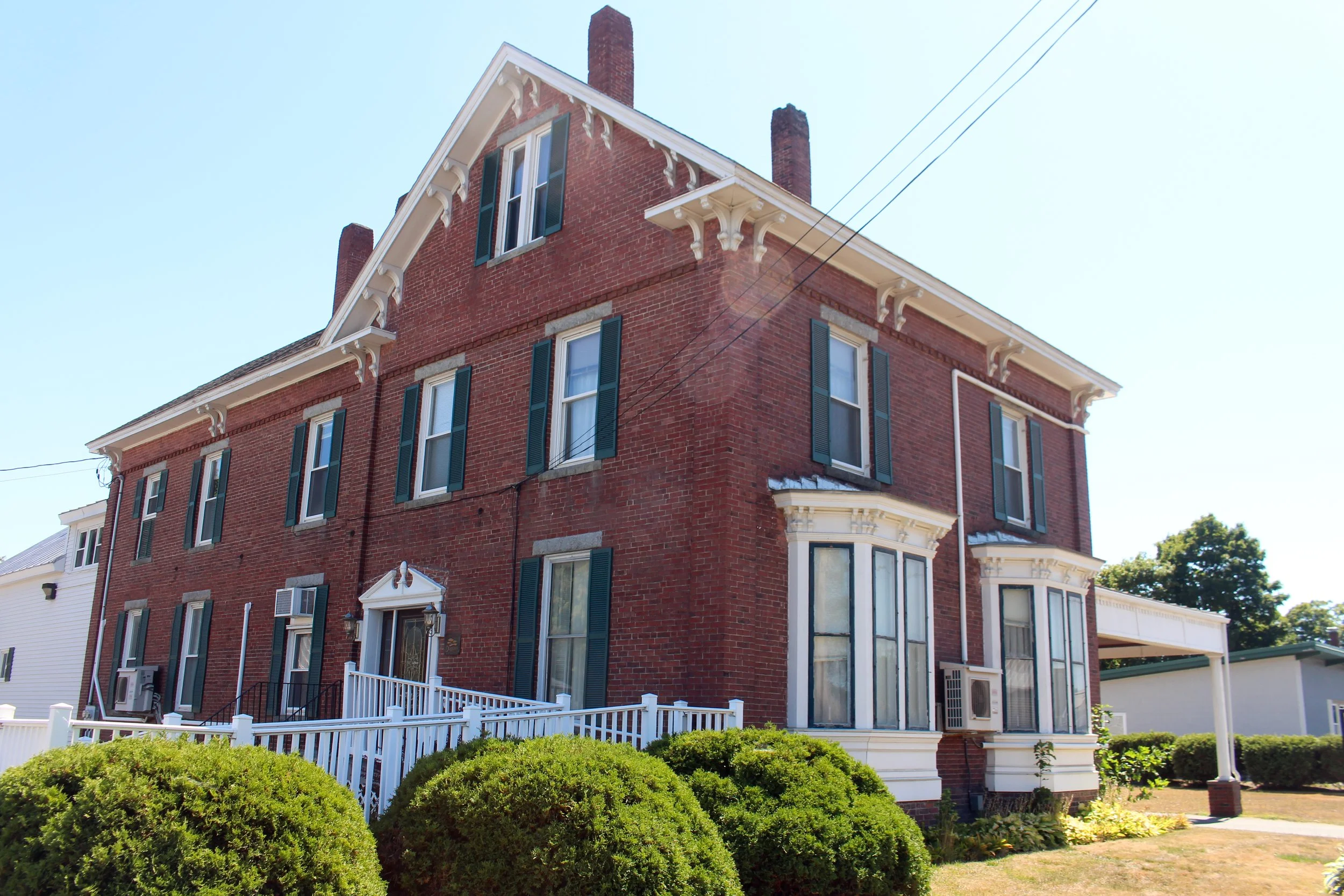 Red brick Victorian-style house with black shutters, white trim, and a fenced front yard with green bushes and a white picket fence.
