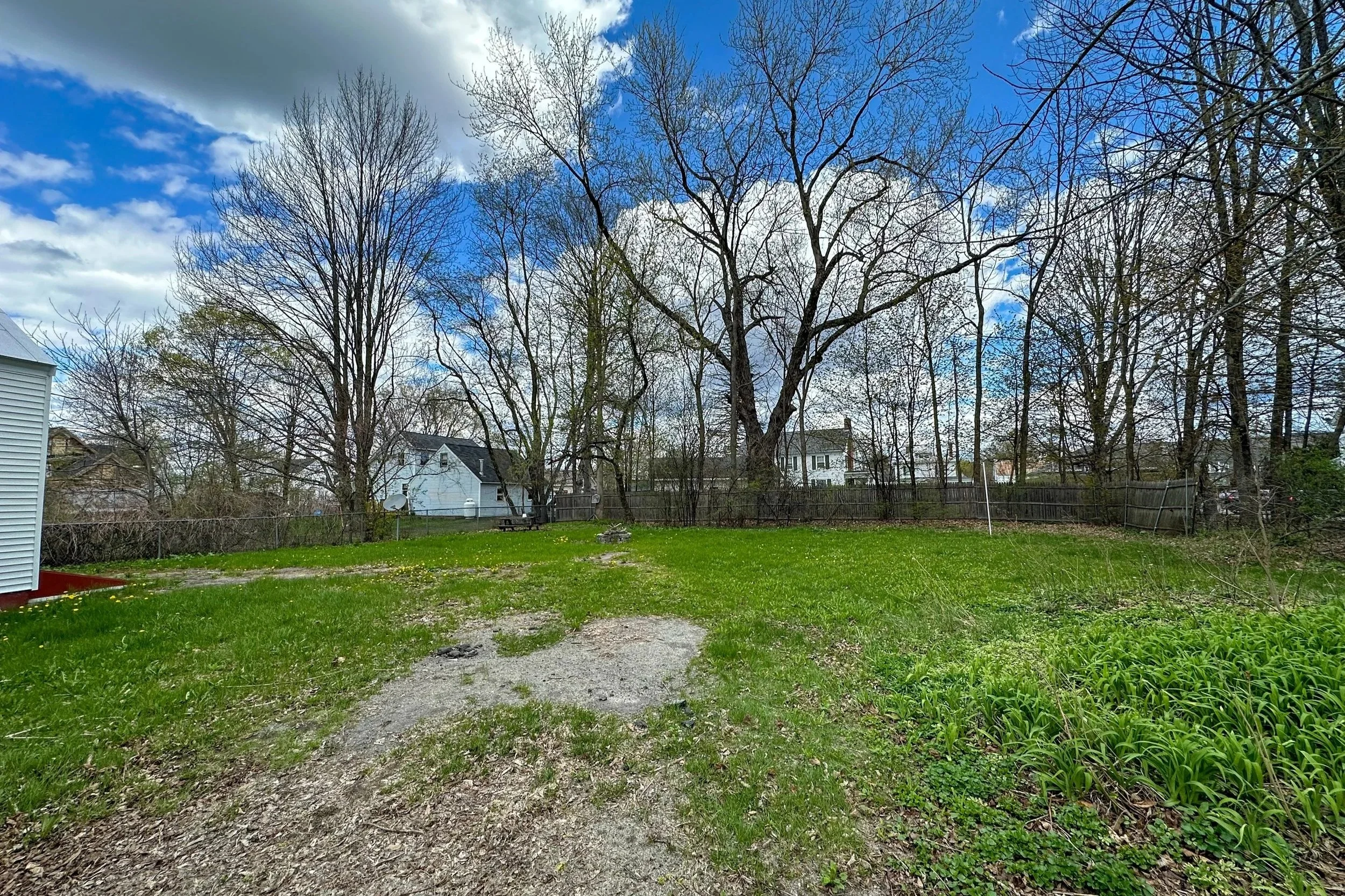 Backyard with green grass, a gravel pathway, leafless trees, a white house, a satellite dish, and a wooden fence, under a partly cloudy sky.