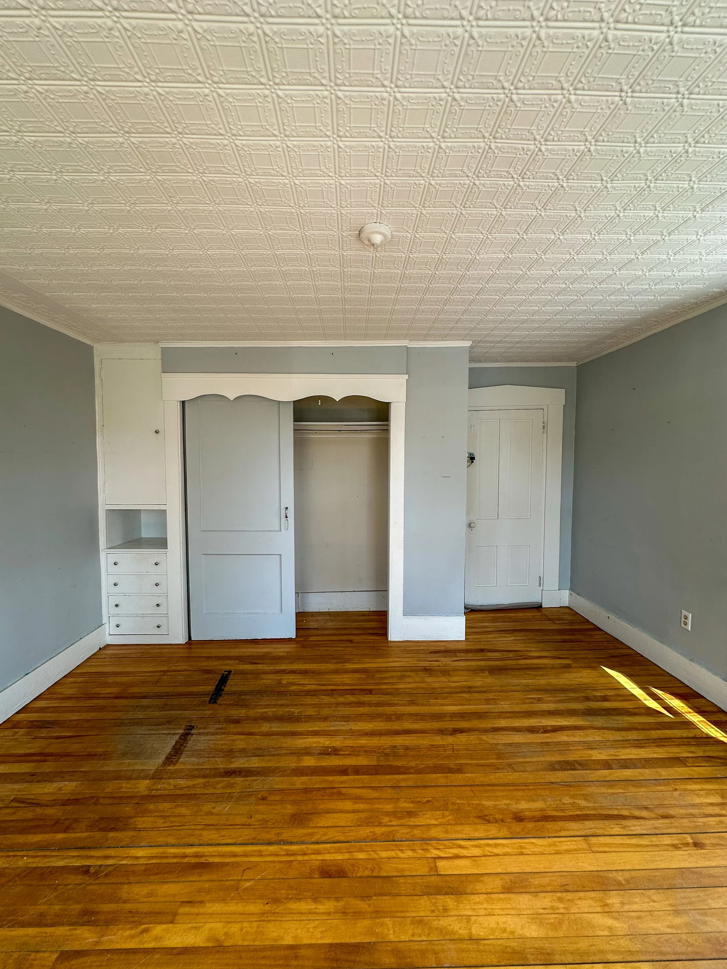 A room with light blue walls, a wooden floor, a white built-in closet with shelves and drawers, and a door. The ceiling has intricate patterned tin tiles, and there is a cord hanging from the ceiling fixture.