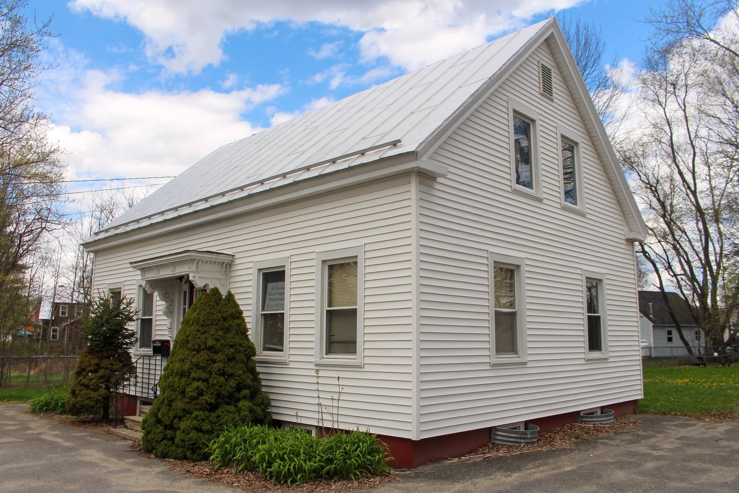 A white two-story house with vinyl siding, multiple windows, and a metal roof. There are two bushes near the front entrance and a small tree. The sky is partly cloudy with patches of blue visible.