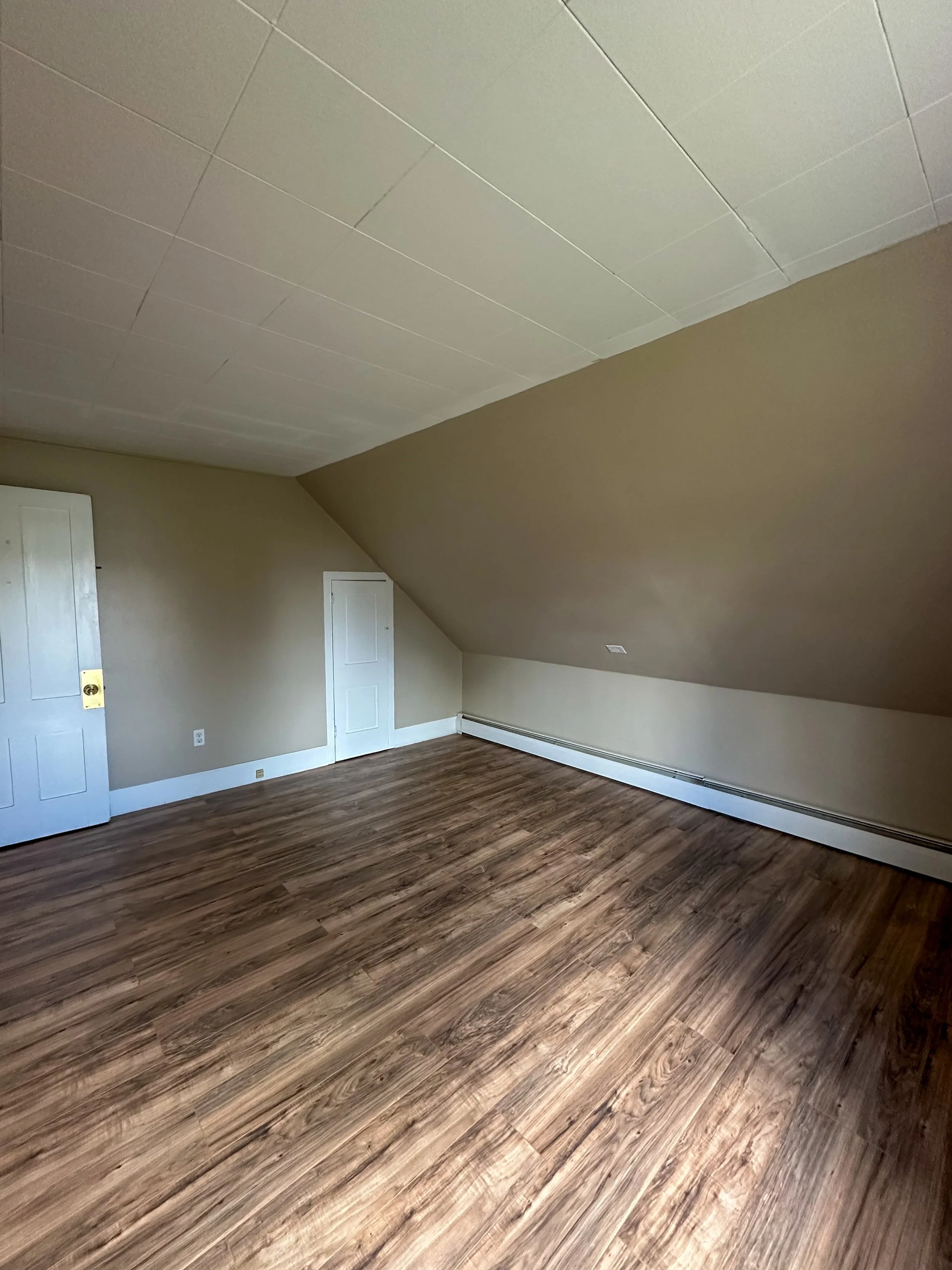 Empty room with beige walls, wood flooring, a white door, two small closet doors, and a sloped ceiling with a vent.