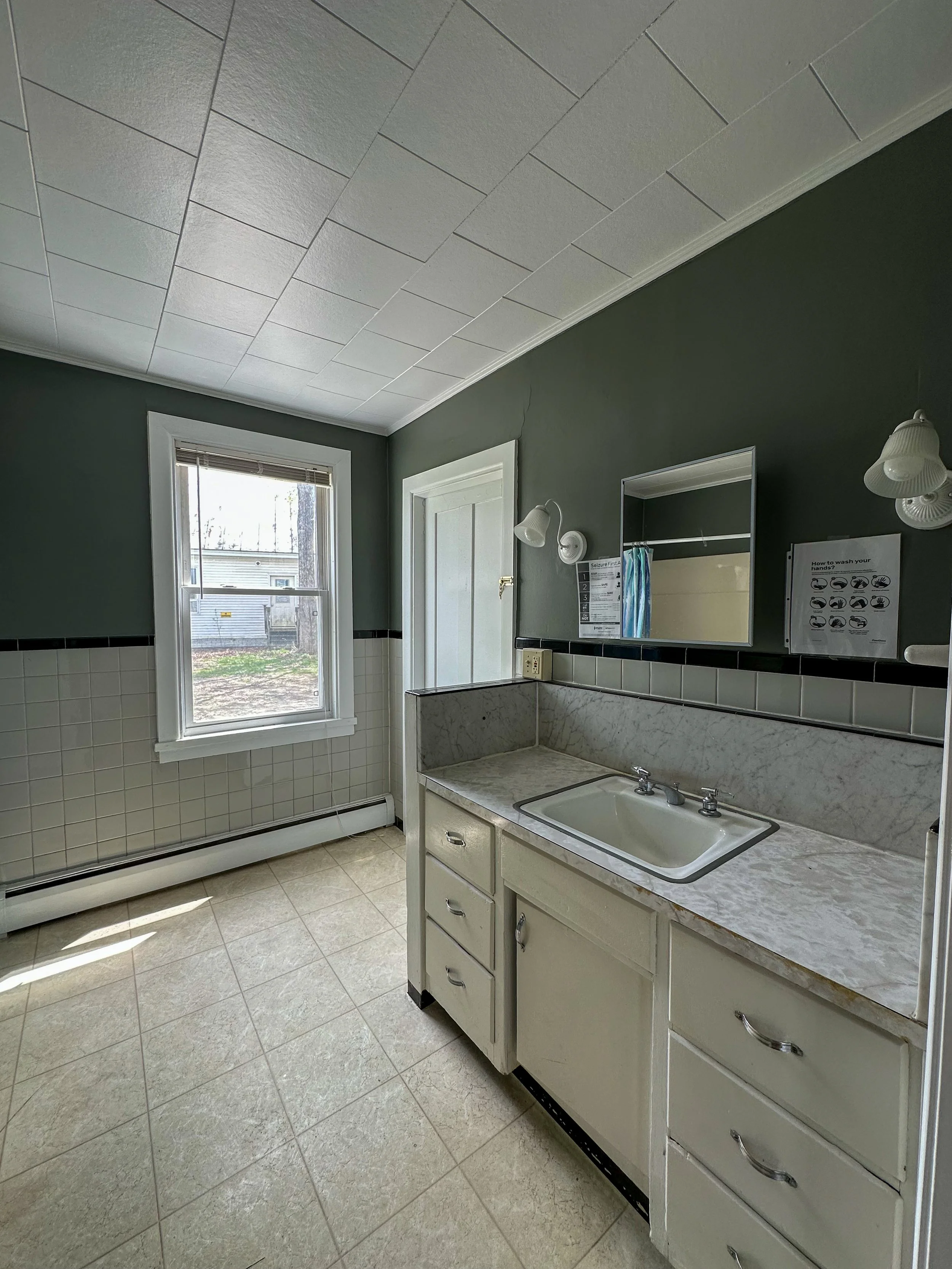 A bathroom with white tiled walls and beige tiled floors, featuring a window, a mirror, and a sink with a cabinet below. There are wall-mounted light fixtures, and the outdoors can be seen through the window.