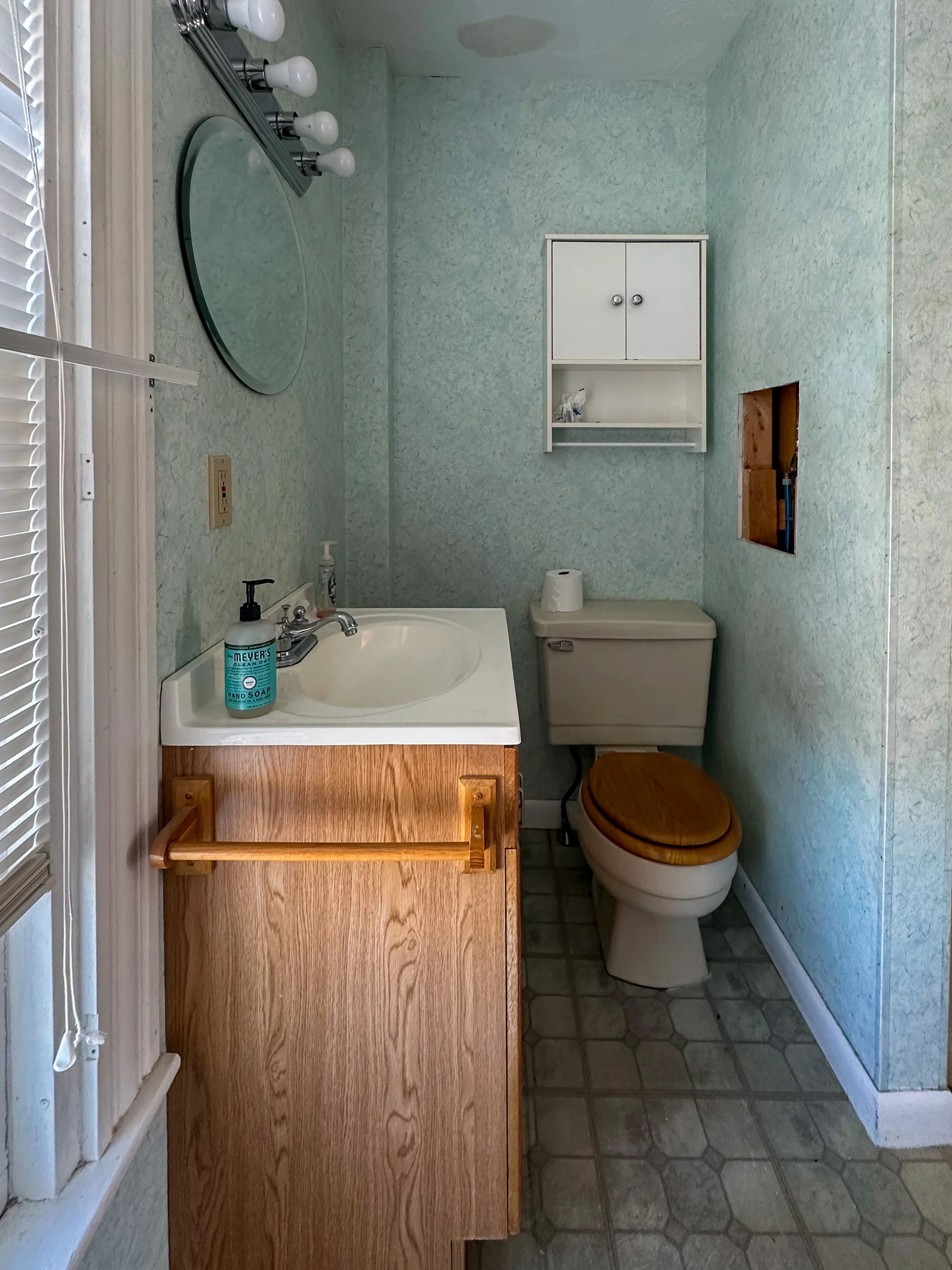 A small bathroom with a white sink and wooden cabinet, a toilet with a wooden seat, a white wall-mounted cabinet, and a hole in the wall revealing plumbing pipes. There are hexagon-patterned floor tiles, a soap dispenser on the sink, a roll of toilet