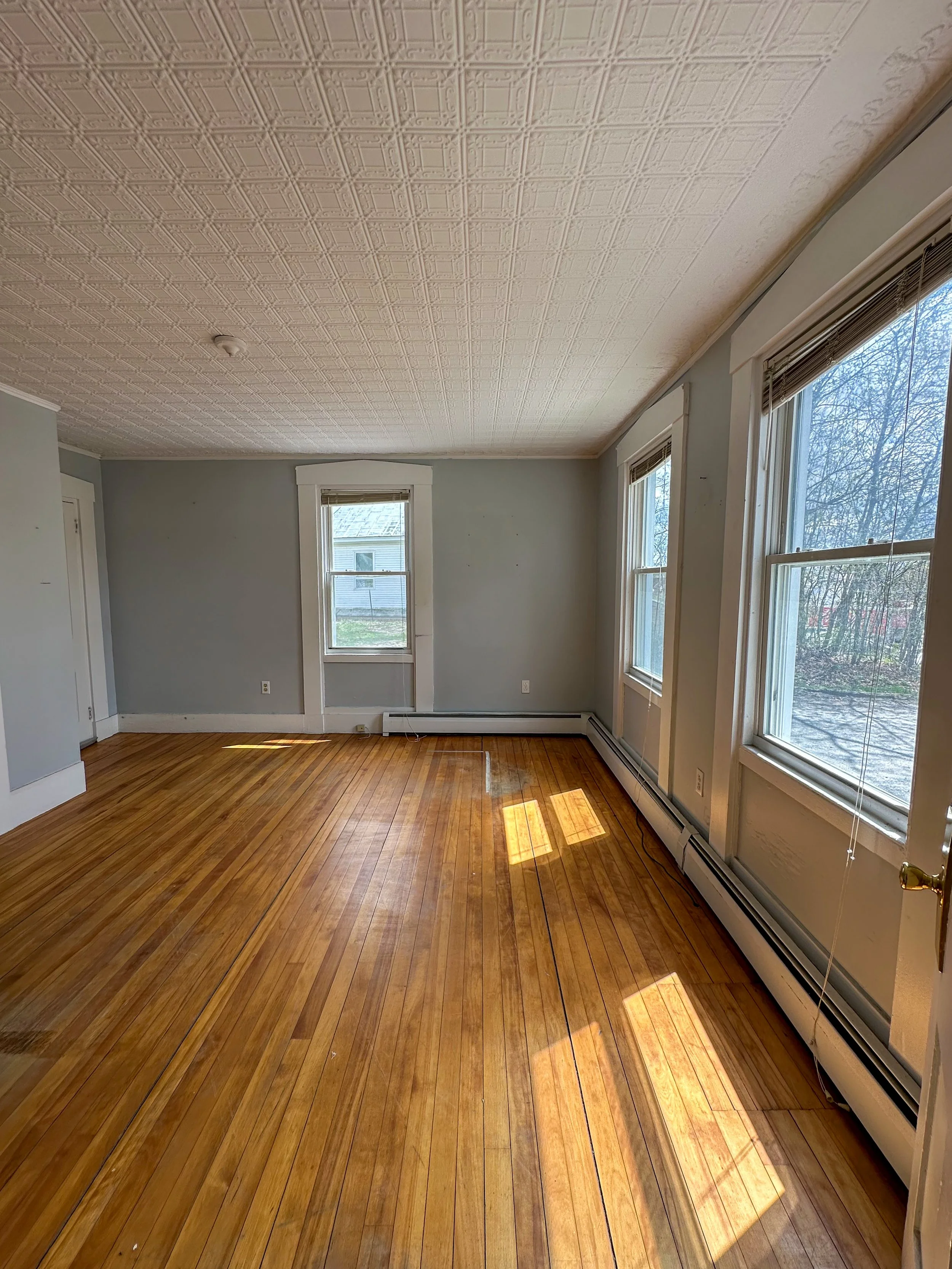 An empty room with beige ceiling tiles, light blue walls, and hardwood floors. There are three windows letting in natural light and a white baseboard heater along the wall.