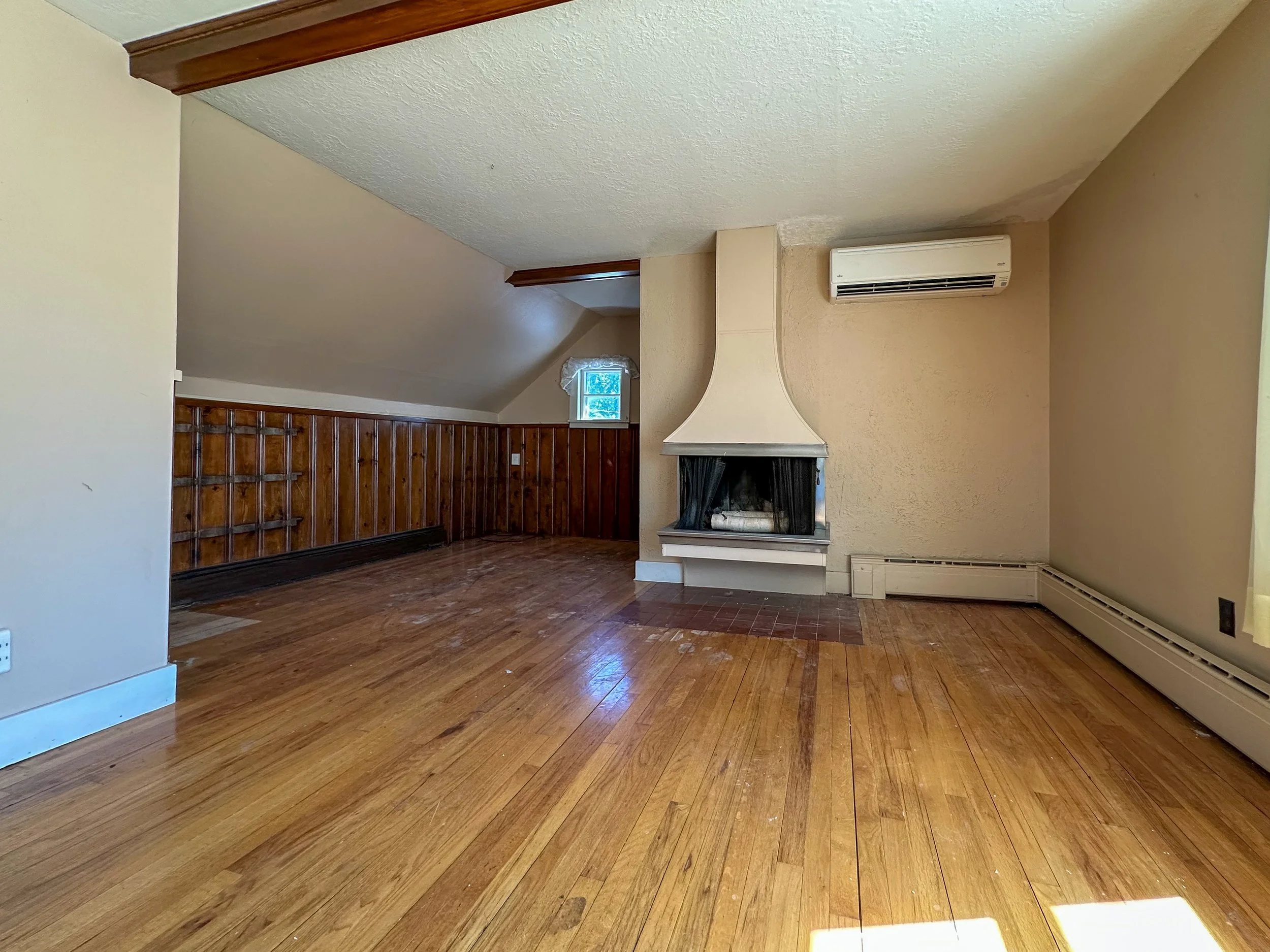 Empty living room with wooden floors, a built-in fireplace with a black metal grate, beige walls, tall baseboard heating, an air conditioning unit, a small window, and wood paneling on one wall.