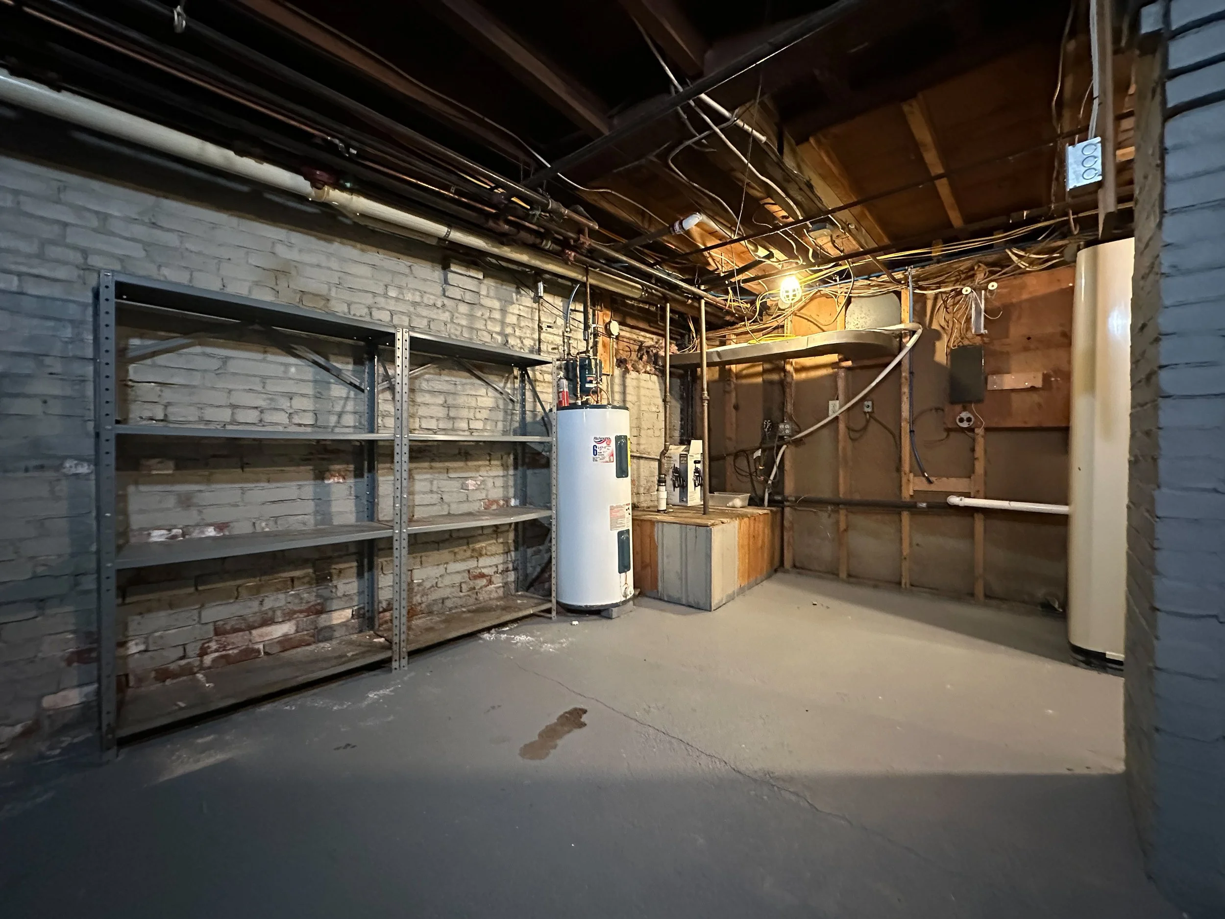 An empty basement or storage room with a brick wall, exposed pipes and wires on the ceiling, a water heater, and a metal shelving unit. The room has a concrete floor and unfinished walls.