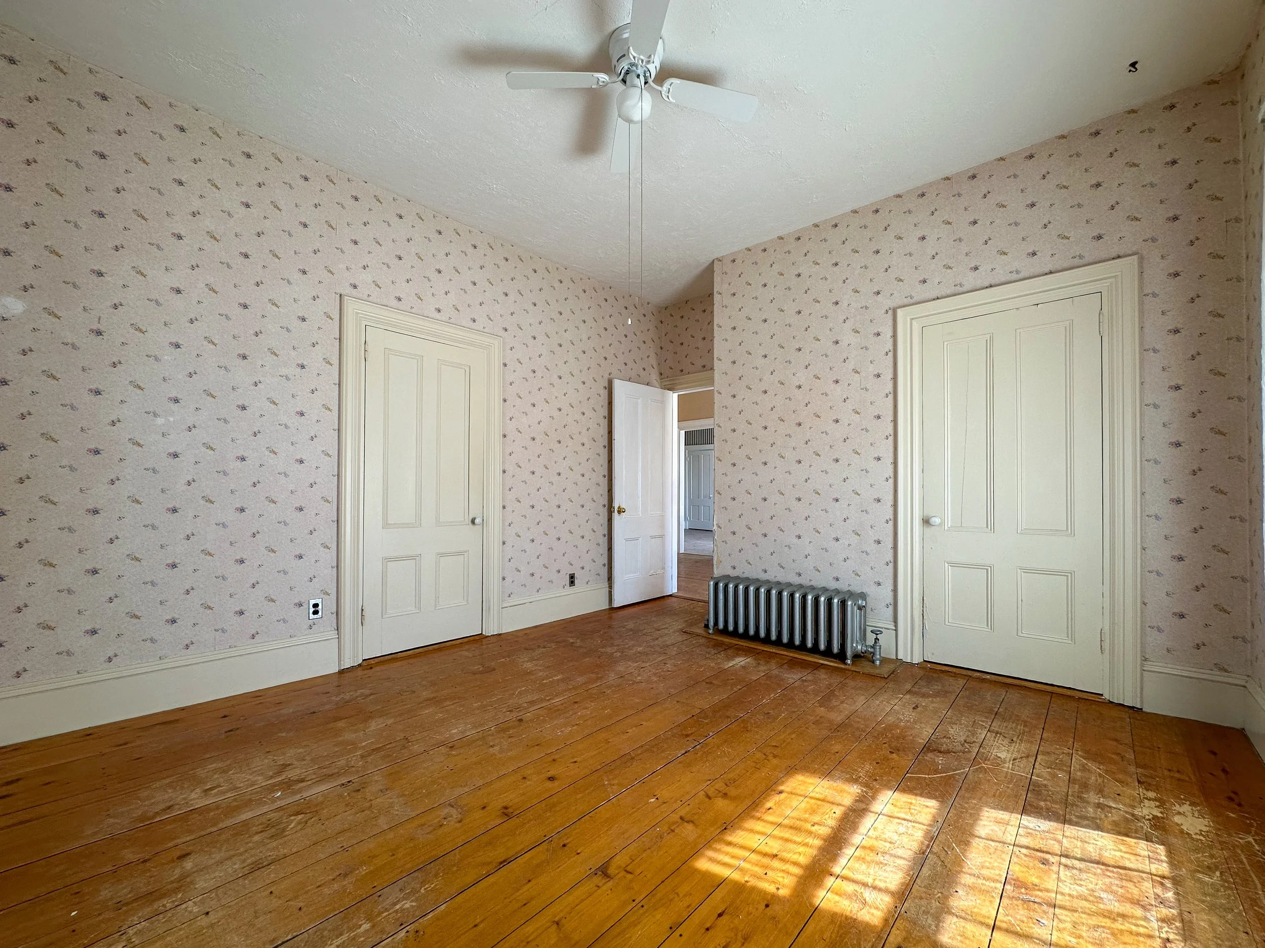 Empty room with hardwood floors, floral wallpaper, a ceiling fan with light, and a radiator heater beside two closed white doors.
