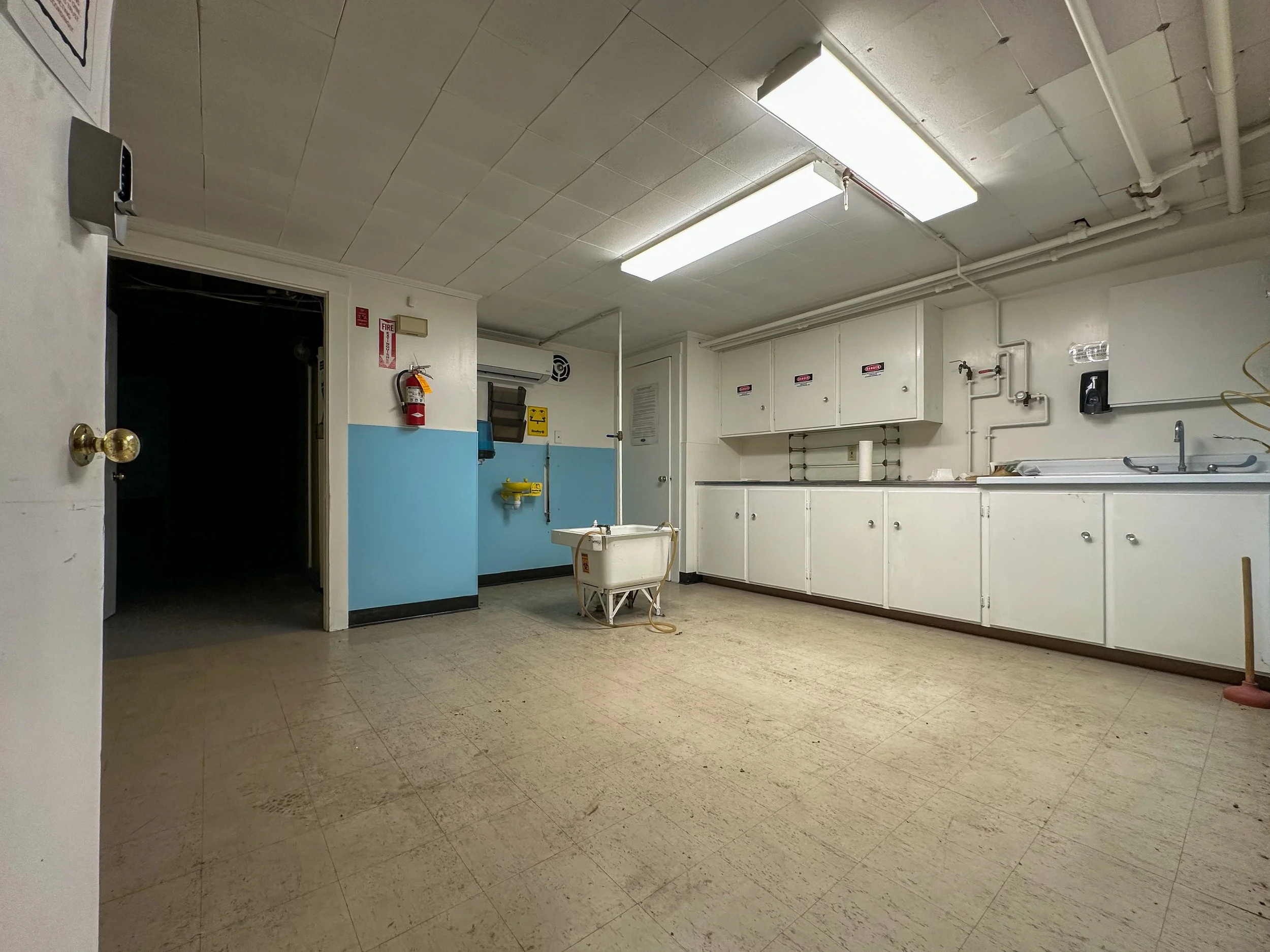 An empty room with white cabinets, a sink, and cleaning supplies, with a tiled ceiling and fluorescent lighting.