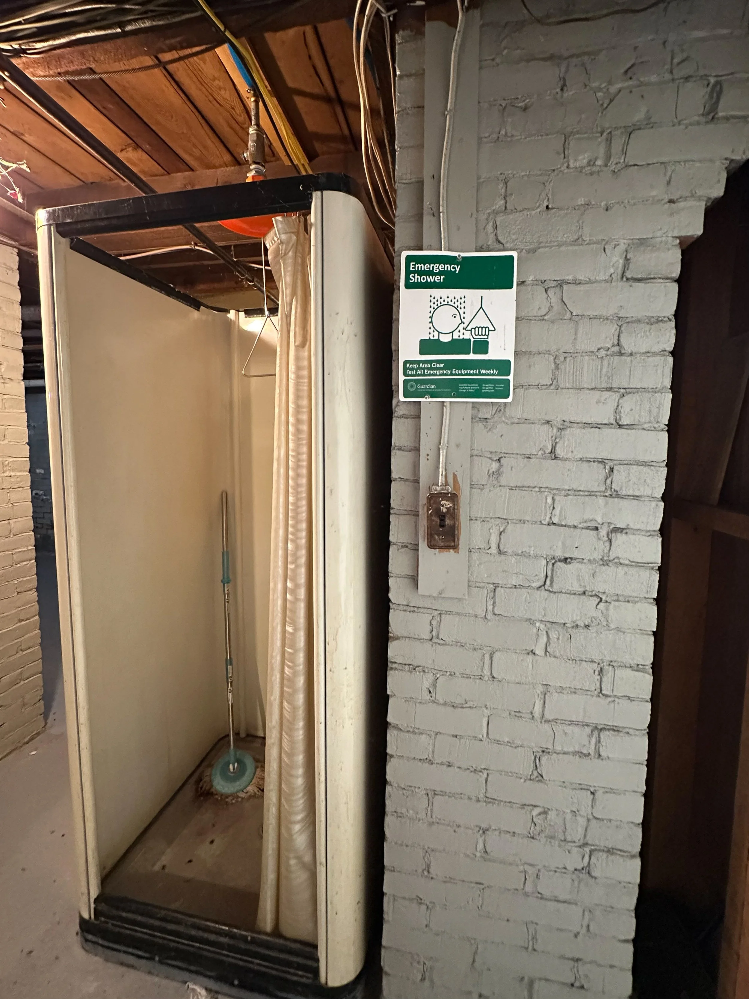 An emergency shower unit with a showerhead and hose, open and unoccupied, located next to a white brick wall with a green emergency shower sign.