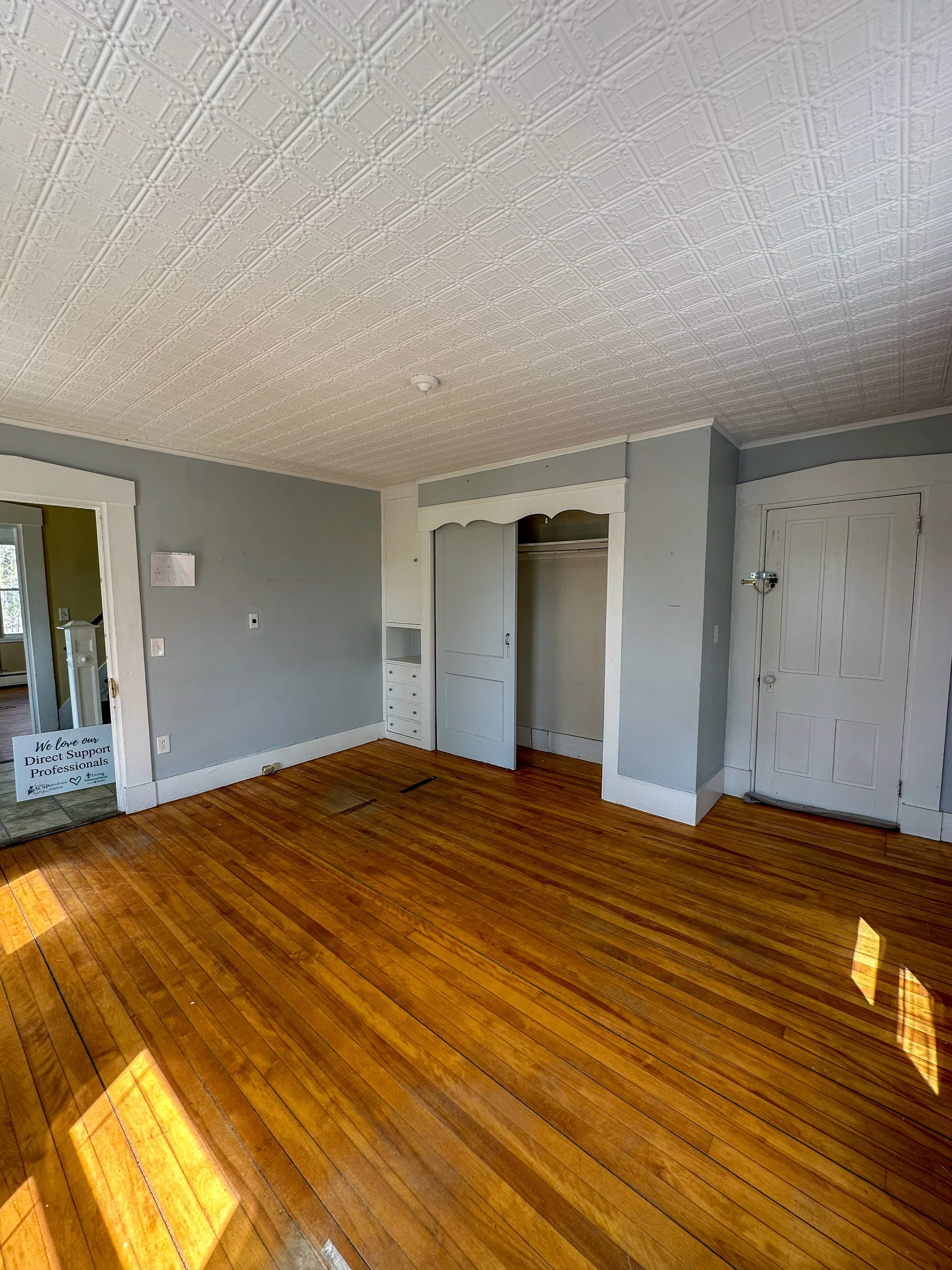 Interior of a room with hardwood floors, light blue walls, a white ceiling with decorative tin tiles, a closet with white doors, and a doorway leading to another room. There is a small sign on the floor near the doorway.