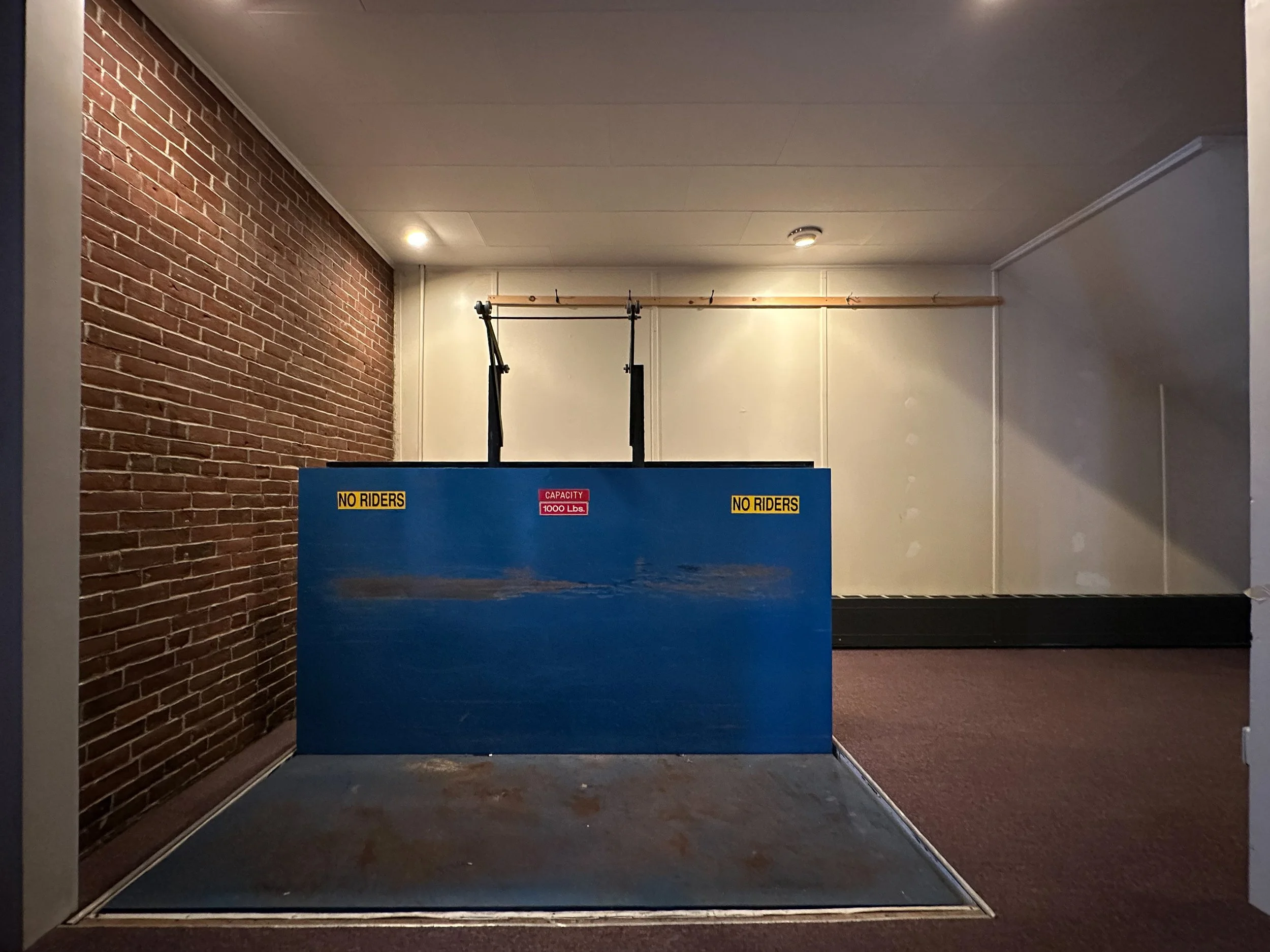 Indoor space with a brick wall on the left and a plain wall on the right, ceiling lights, and a large blue structure with warning signs for no riders and a weight capacity of 1000 lbs, possibly part of an amusement or gym equipment.