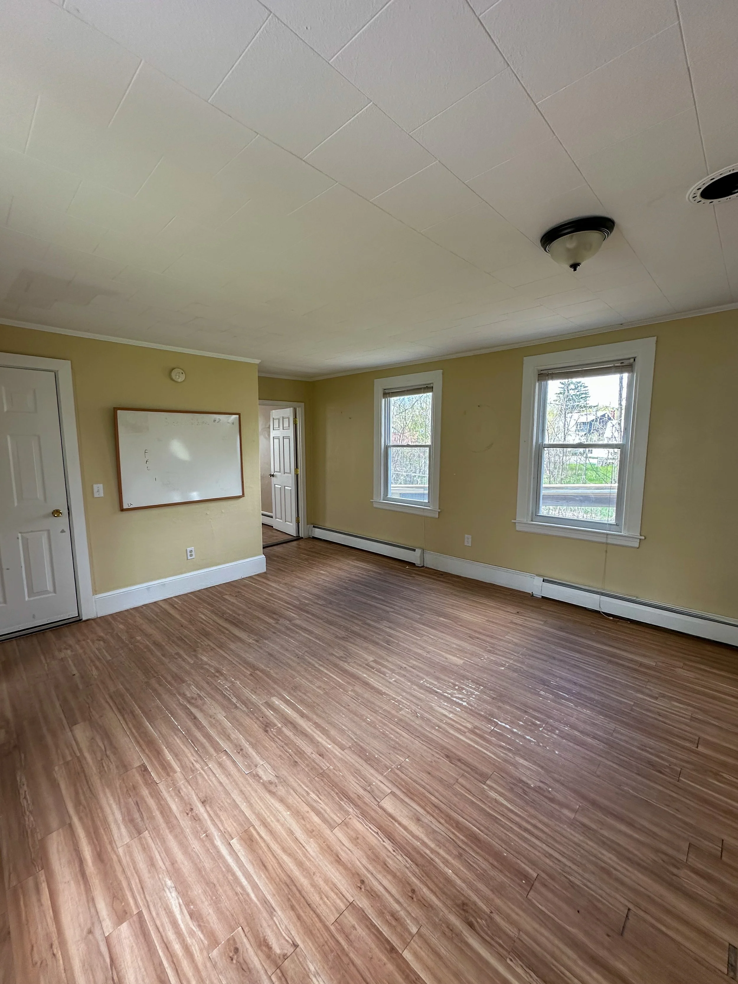 Empty room with wood-look flooring, beige walls, three windows with view of trees and neighboring house, white trim, white baseboard heater, white door, and whiteboard on wall.