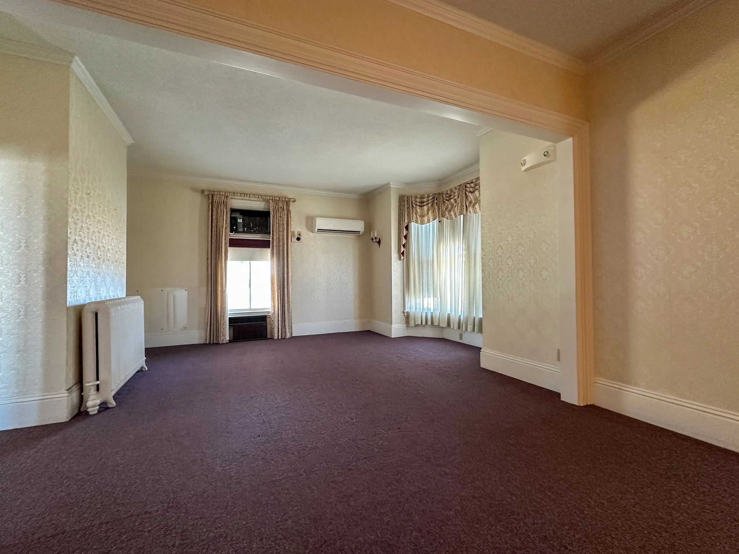 Interior room with beige walls, patterned curtains on windows, dark carpeting, and a white radiator.