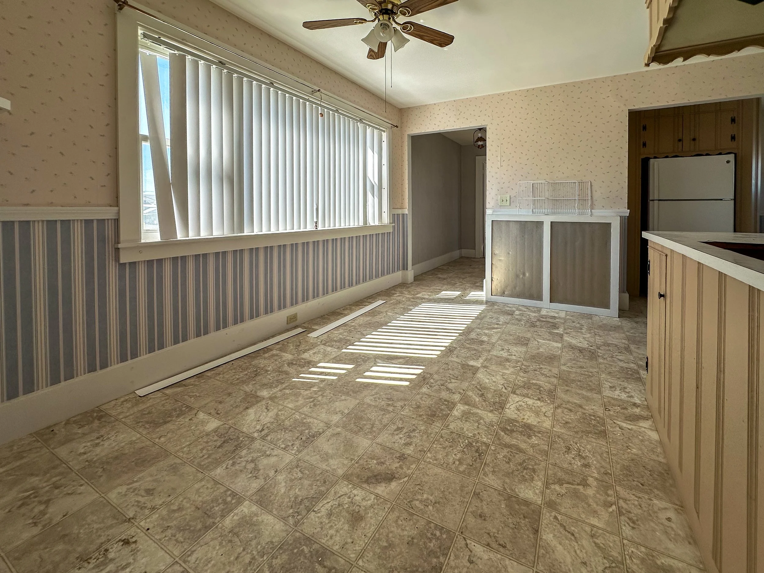 Sunlight streams into a room through vertical blinds, casting shadows on beige tiled flooring. The room has wallpaper with small patterns, a ceiling fan with wooden blades, and a corner kitchen with a white refrigerator and wooden cabinets.