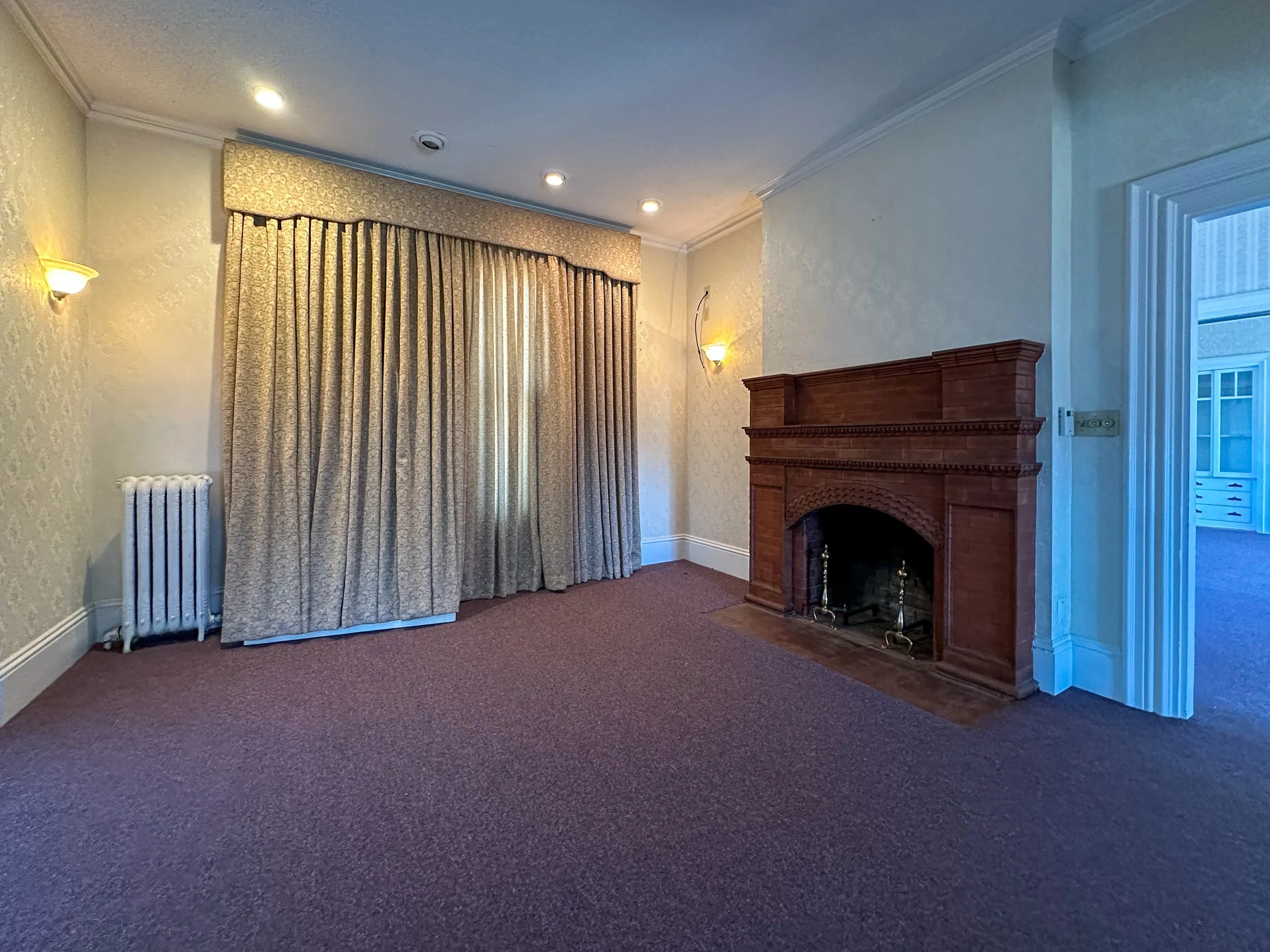 Living room with beige patterned curtains, wooden fireplace, beige textured walls, wall-mounted lights, brown carpet, and a potted radiator.