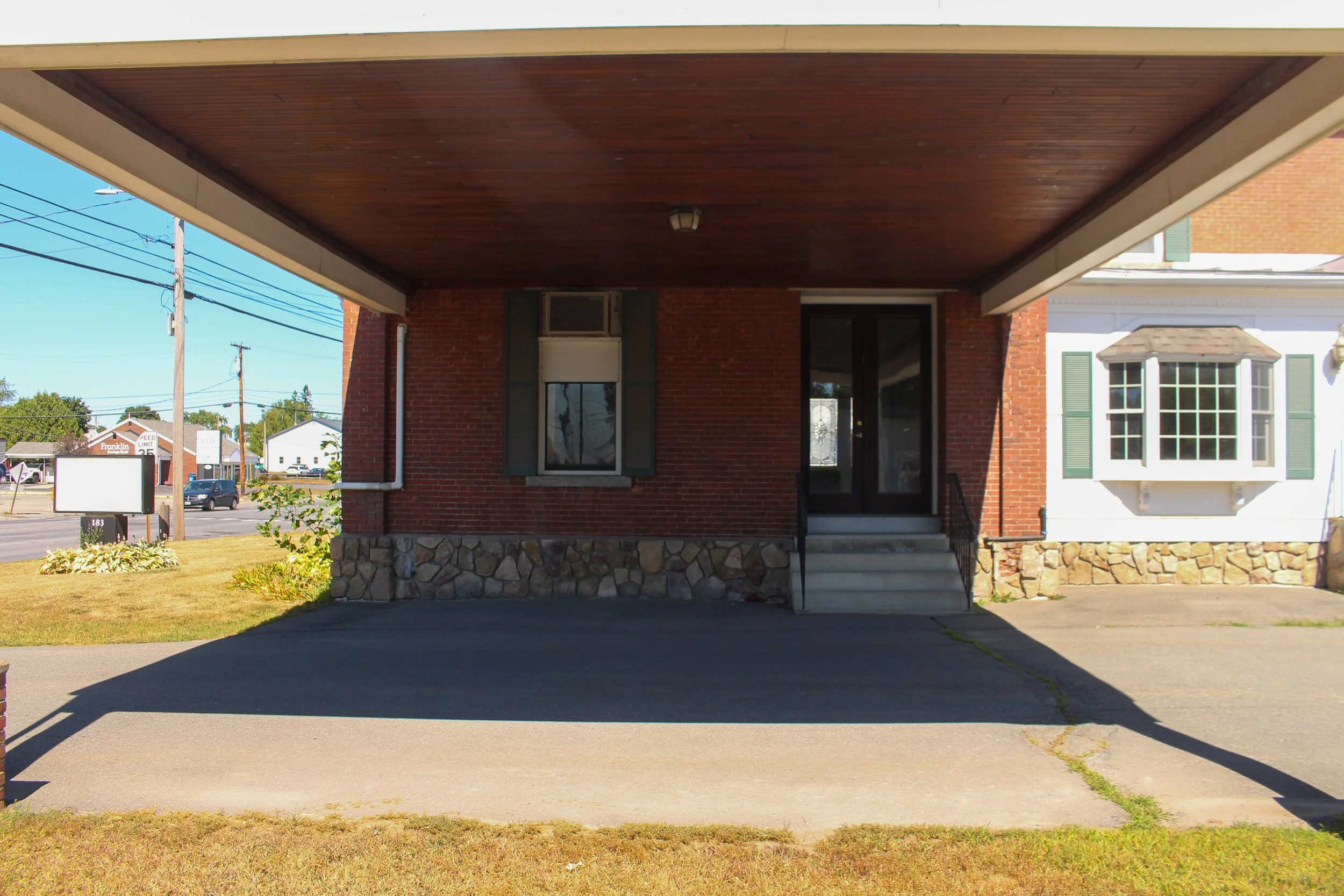 Front view of a brick house with a covered porch, stone steps, and a glass door. A window with shutters is visible on the right side. The yard has a small garden bed and grass, with a street and power lines in the background.