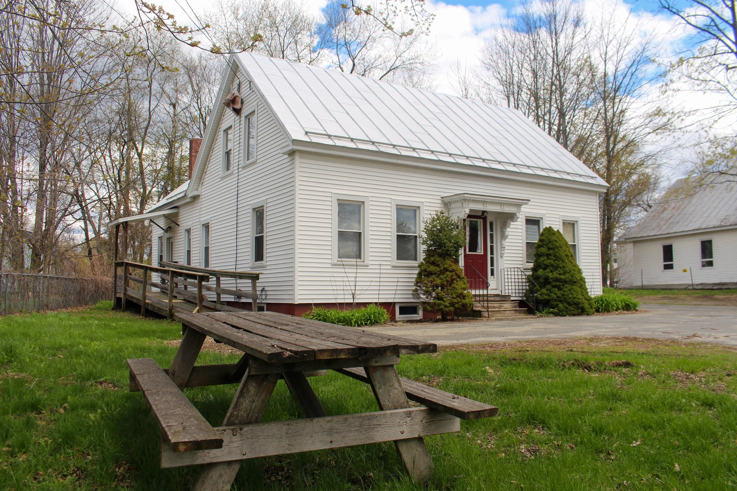 White wooden house with a metal roof, front porch, and small trees and bushes in front, surrounded by trees on a cloudy day.