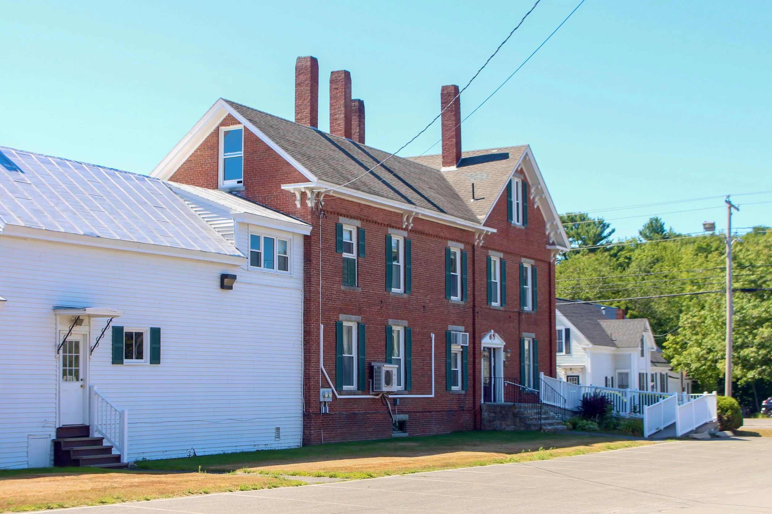 Residential street view featuring a white house with green shutters and a red brick building with multiple chimneys, set against a blue sky with utility wires and trees in the background.