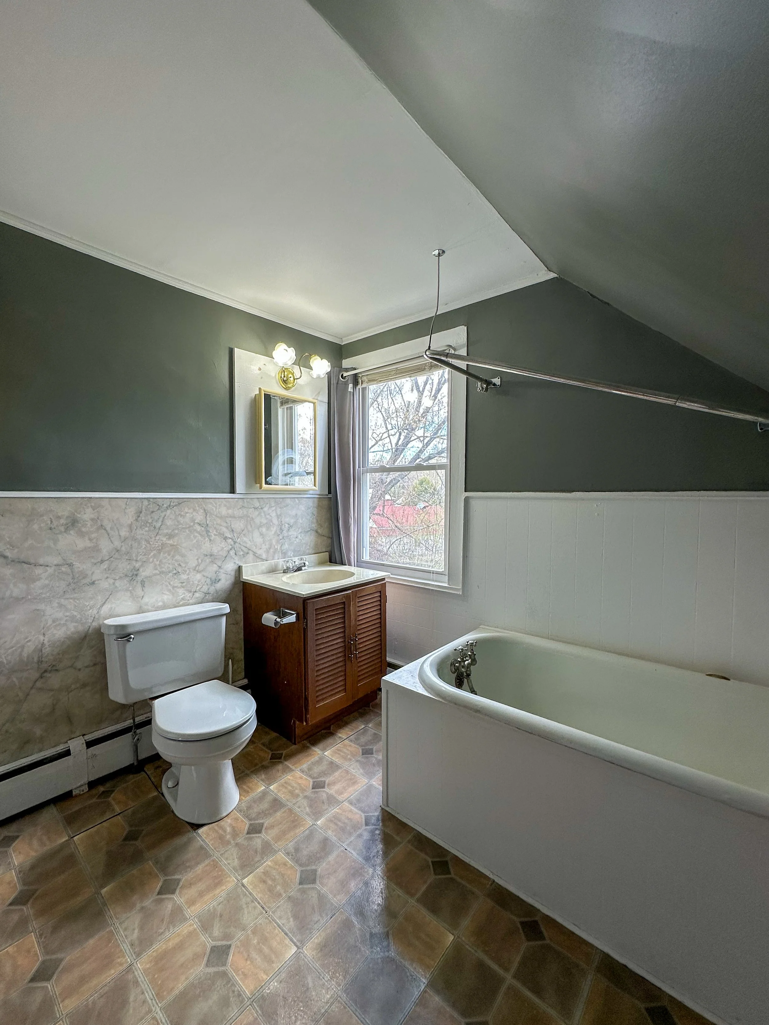 Bathroom with green walls, window, bathtub, toilet, small wooden vanity with sink, mirror, light fixture, and patterned tile floor.