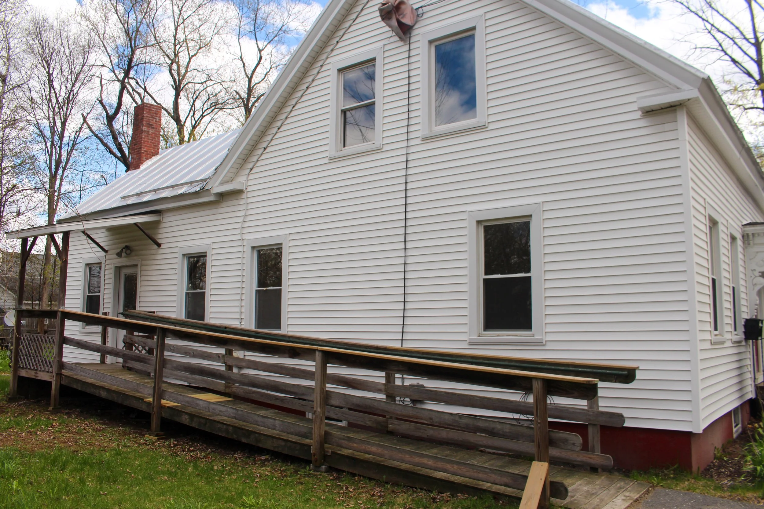 Back of a white house with vinyl siding, multiple windows, a brick chimney, and a metal roof. A wooden deck with a railing is under construction in the yard, with trees in the background.