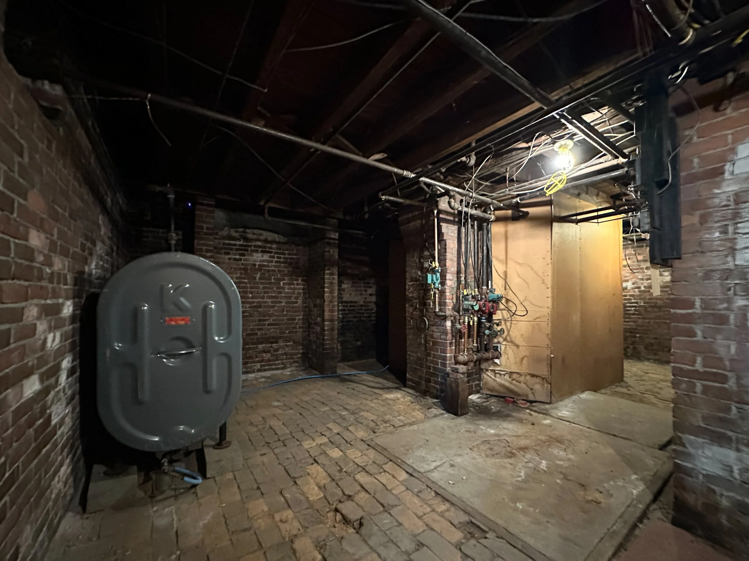 An unfinished brick room with exposed pipes, electrical wires, and a single light bulb hanging from the ceiling.