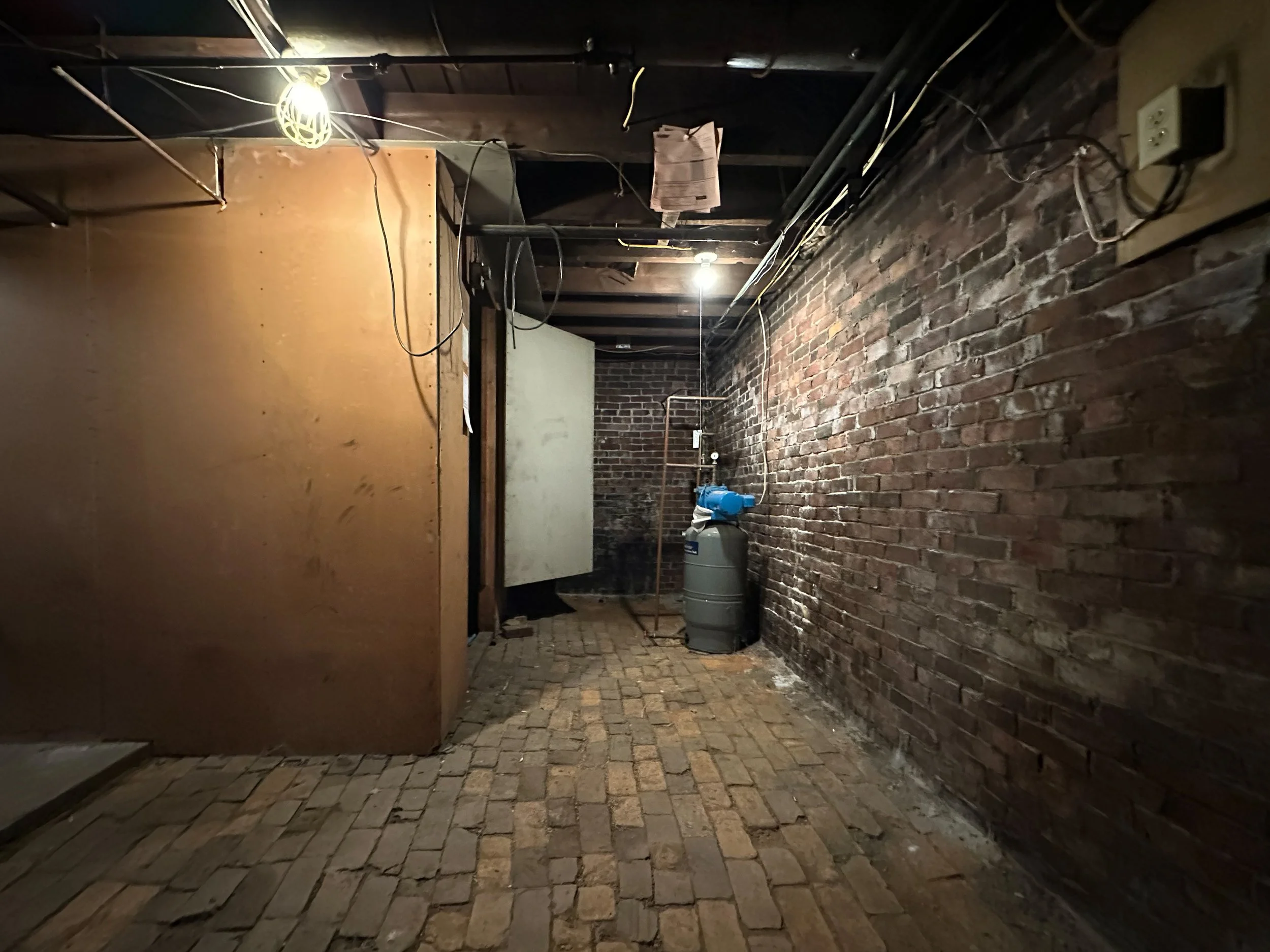 A dimly lit basement or utility room with brick walls and a cobblestone floor. There is a large grey water heater or boiler in the corner, and exposed pipes and wiring on the ceiling. A yellowish panel partially covers one wall, with wires hanging ne