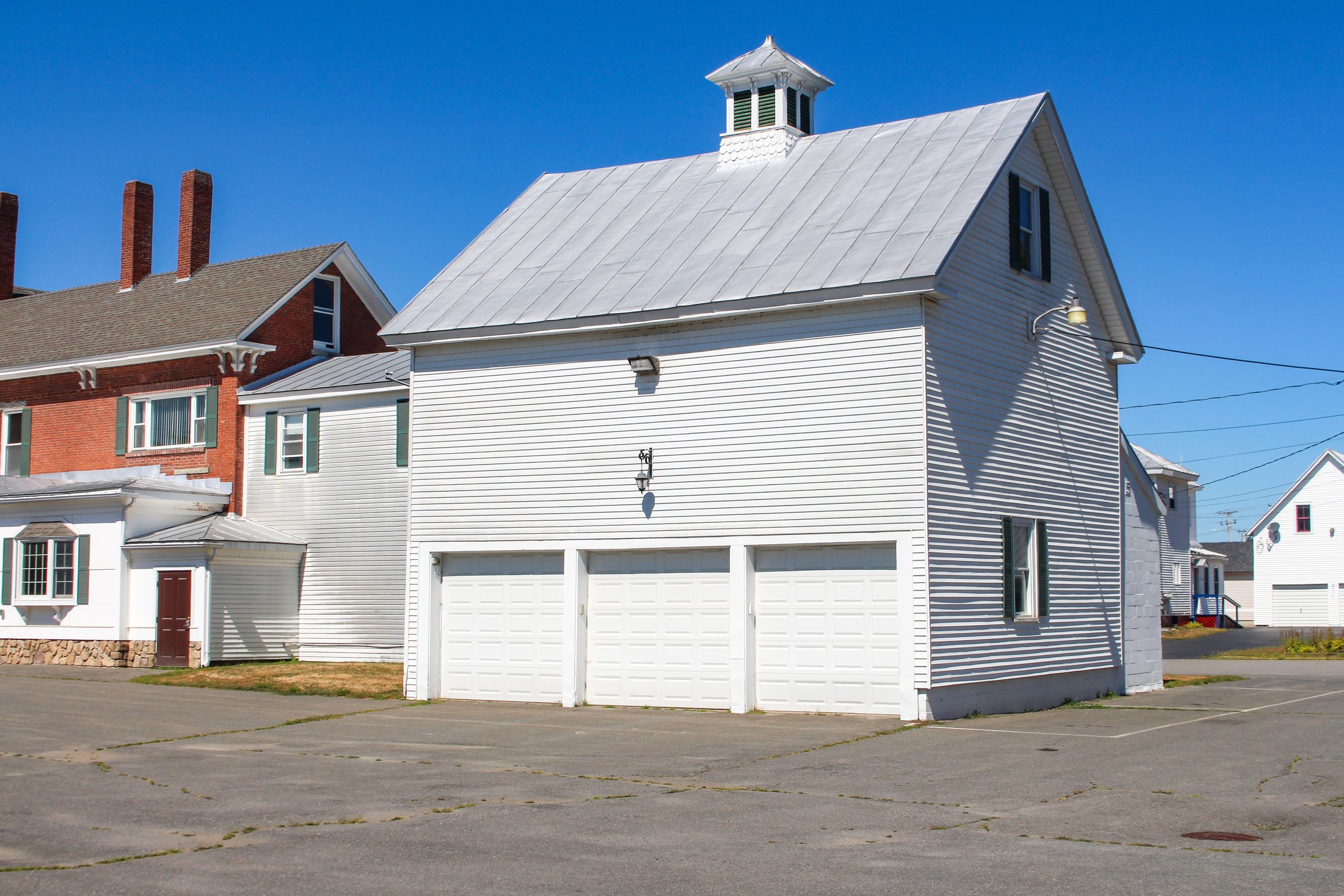 White house with a metal roof, garage door, and a small attic window, situated on a paved lot with other houses nearby, under a clear blue sky.
