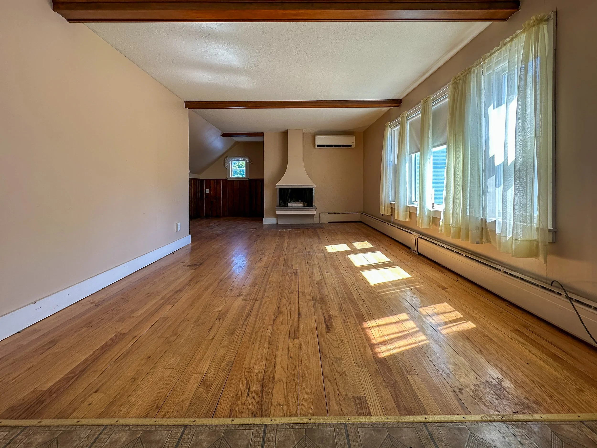 An empty living room with hardwood floors, cream-colored walls, a small window, beige curtains, a white fireplace, an air conditioning unit, and multiple windows letting in natural light.