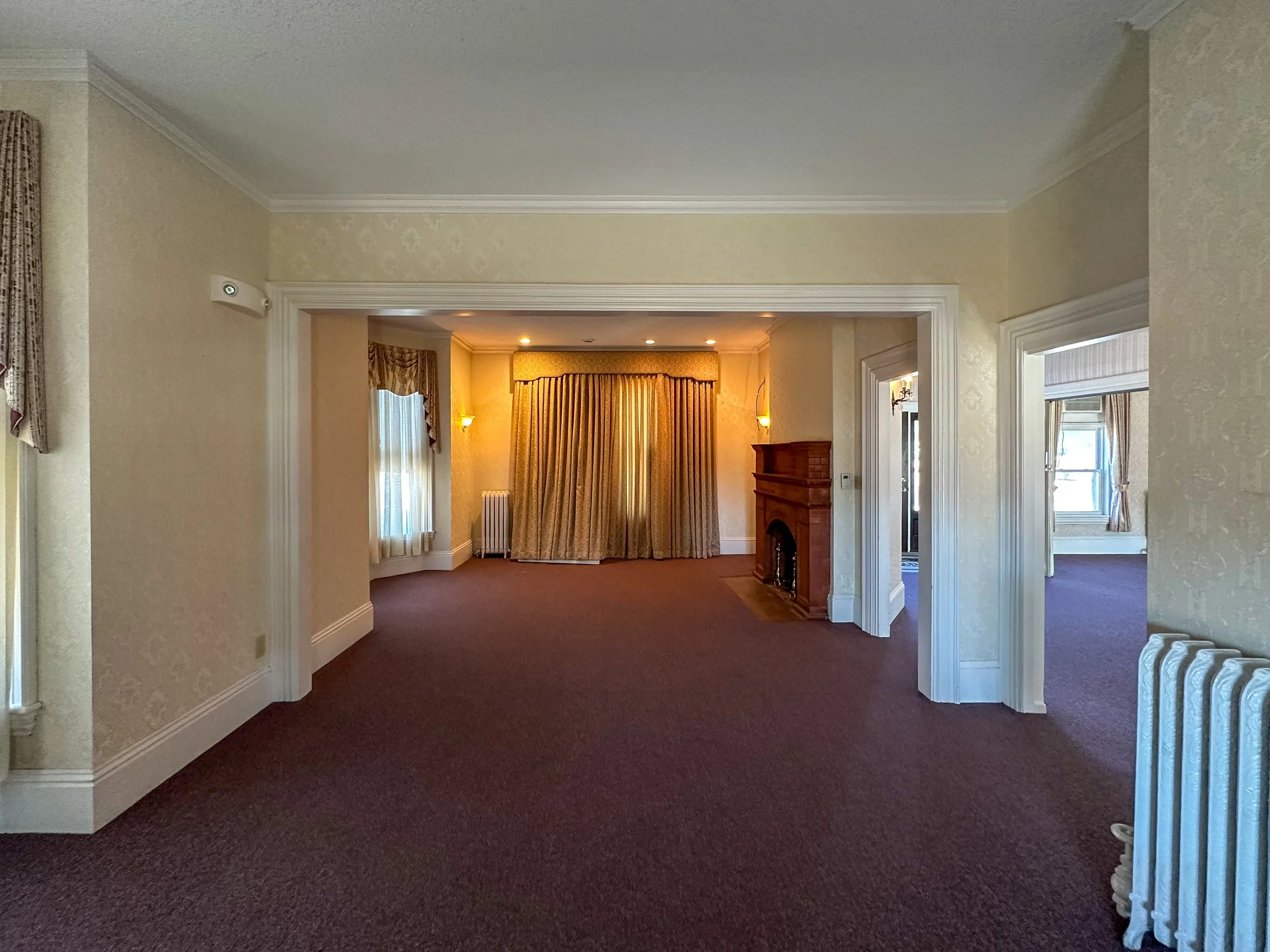 Interior view of a spacious living area with a view into an adjacent smaller room, featuring beige walls, white crown molding, dark red carpet, windows with curtains, a wooden fireplace, and recessed lighting.