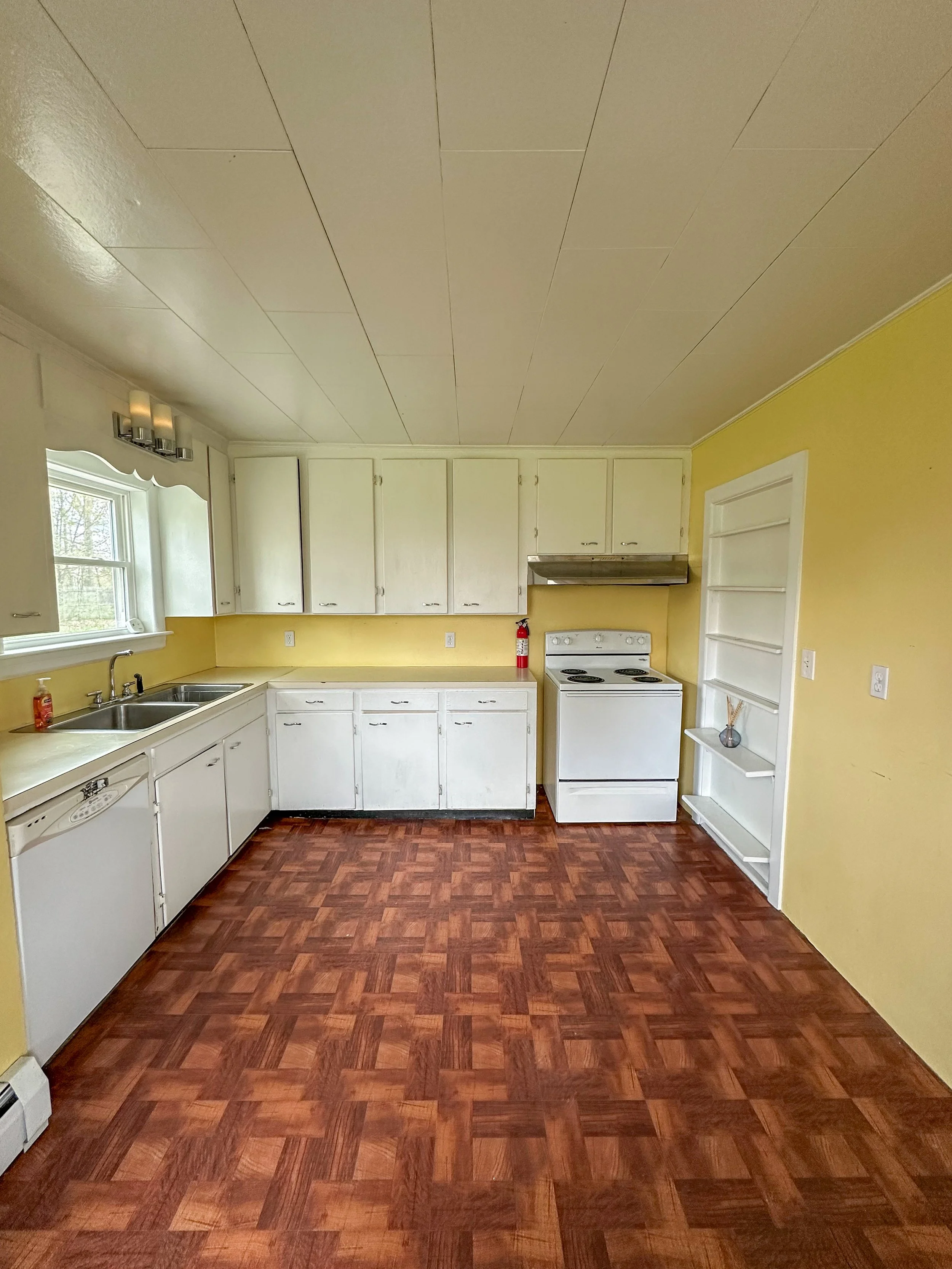 A kitchen with white cabinets and a yellow wall, a window above the sink, a stove, and a decorative shelf. The floor has a red-brown wood pattern.