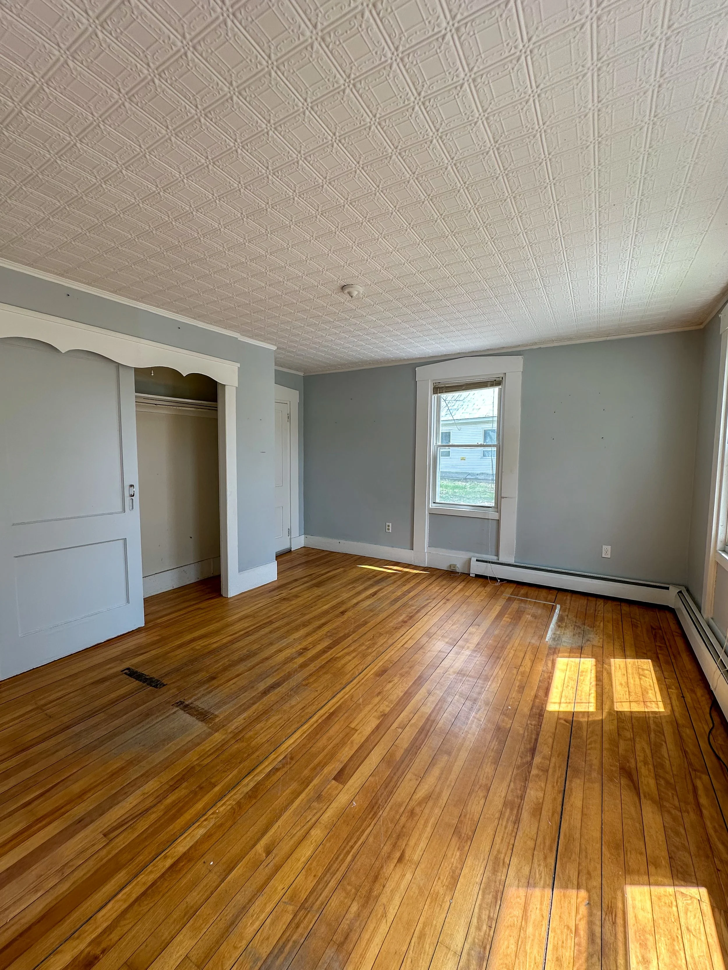 Empty room with hardwood floors, light green walls, large window, and ceiling with decorative tiles.