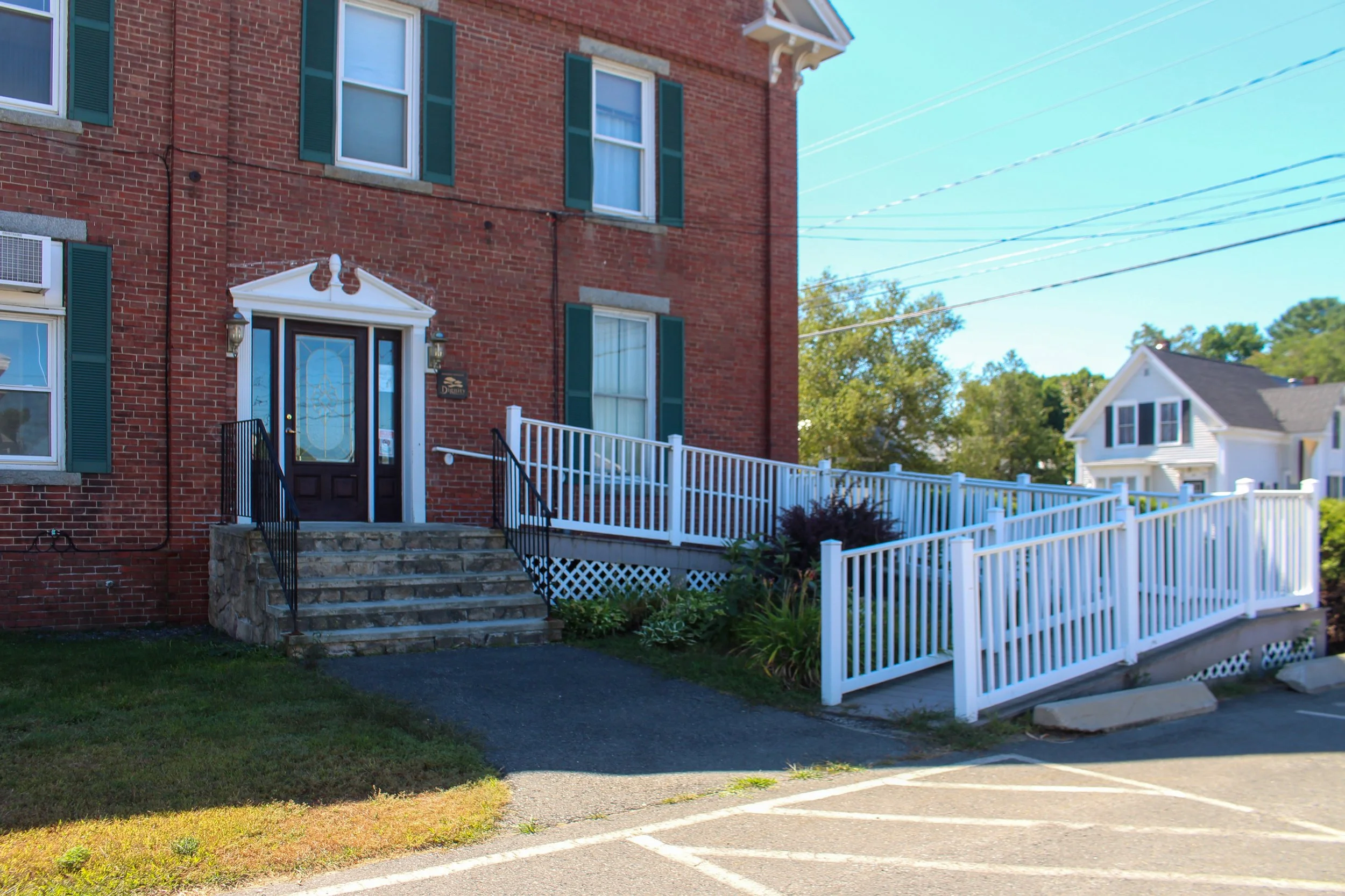 A brick residential building with multiple green shutters and a front door with steps leading up to it. The building has a white fence and surrounding greenery, with a blue sky overhead.