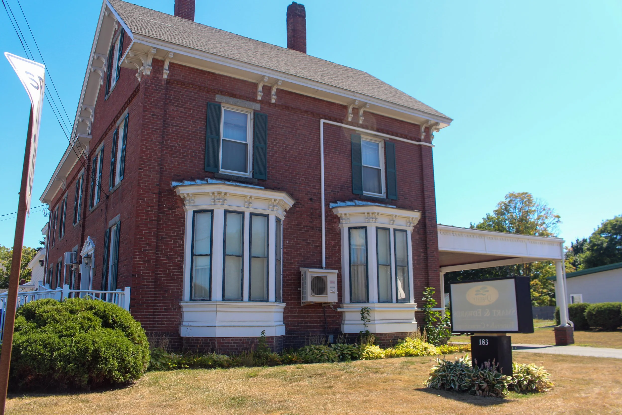 A red brick Victorian-style house with bay windows, dark green shutters, white decorative trim, and a steep shingle roof, with a manicured lawn and small bushes in front.