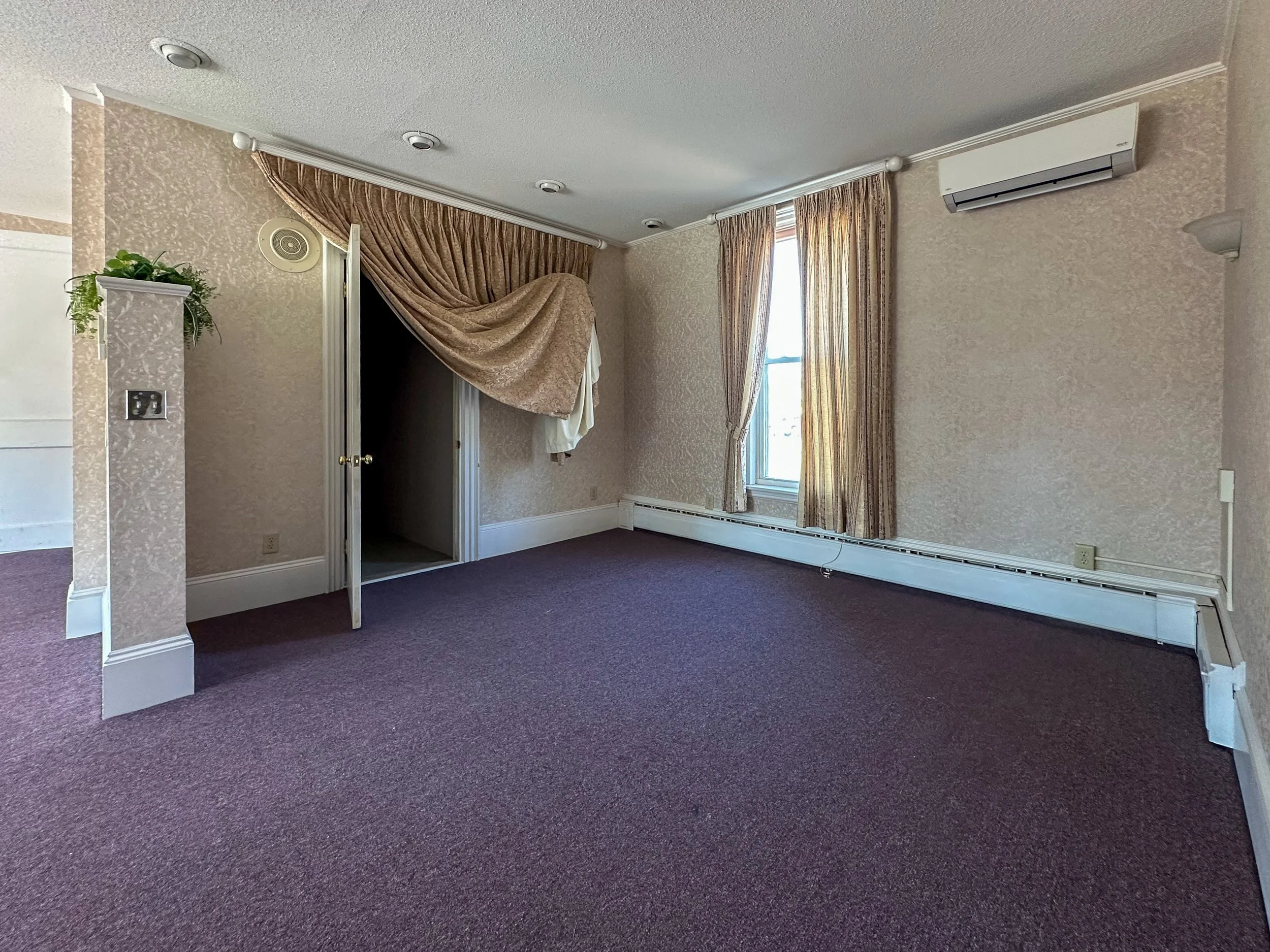Interior view of a room with beige patterned wallpaper, large windows with beige curtains, and purple carpet flooring. There is an open closet with a draped curtain, a plant on a shelf, and an air conditioning unit mounted on the wall.