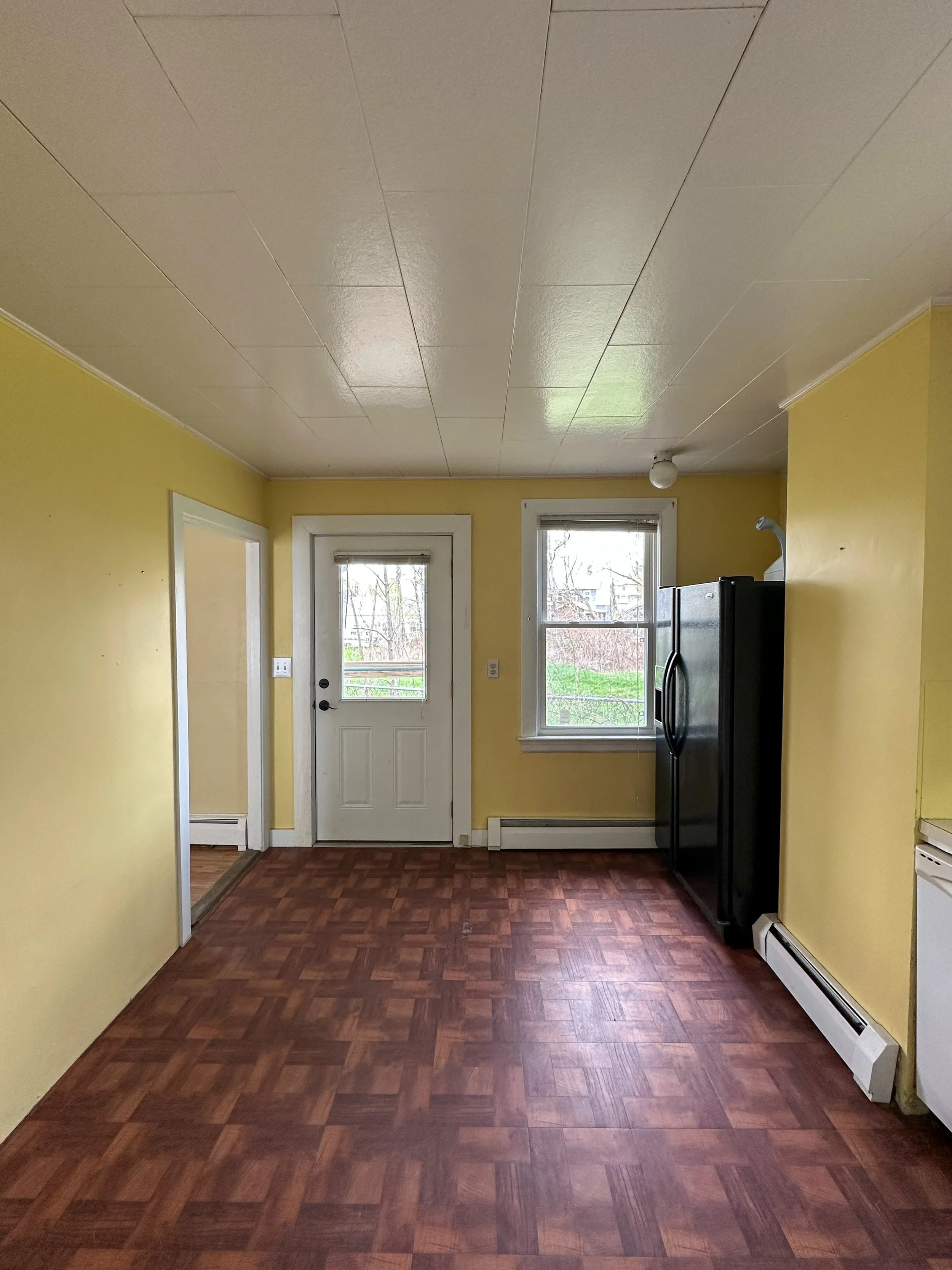 Empty kitchen with yellow walls, wooden floor, two windows, a door, and a black refrigerator.