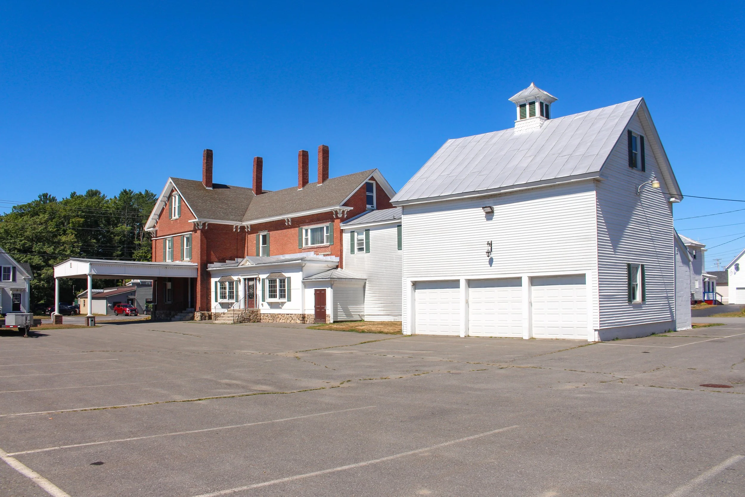 A large paved parking lot with white lines and no cars, adjacent to a white building with a garage and a blacklamps. In the background, there is a two-story red brick house with a prominent roof, four chimneys, and a small porch, along with trees and